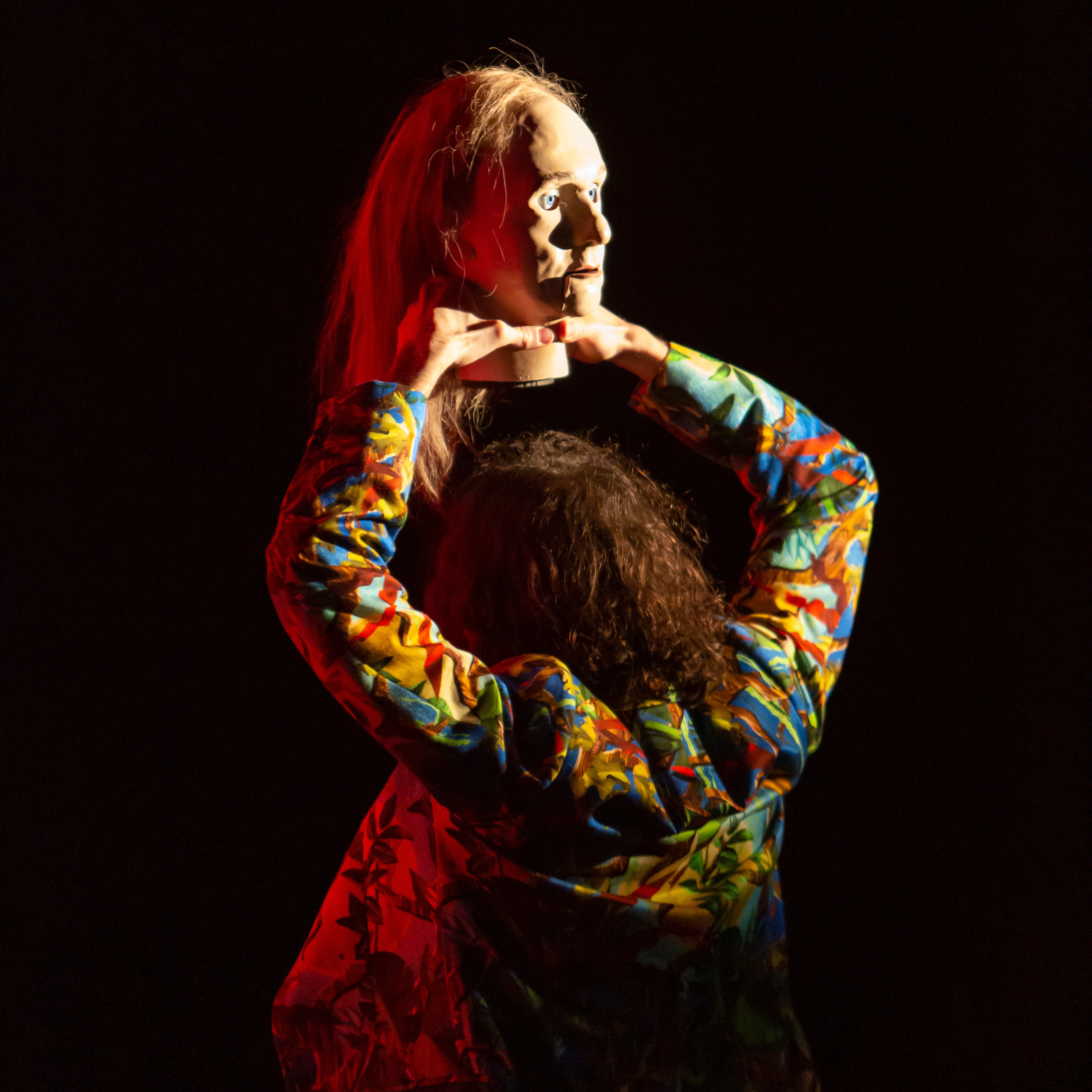 Diederik Peeters in einem sehr bunten Anzug dreht uns den Rücken zu. Diederik hält eine Art lebensgrossen Puppenkopf mit langen Haaren über seinen eigenen Kopf.