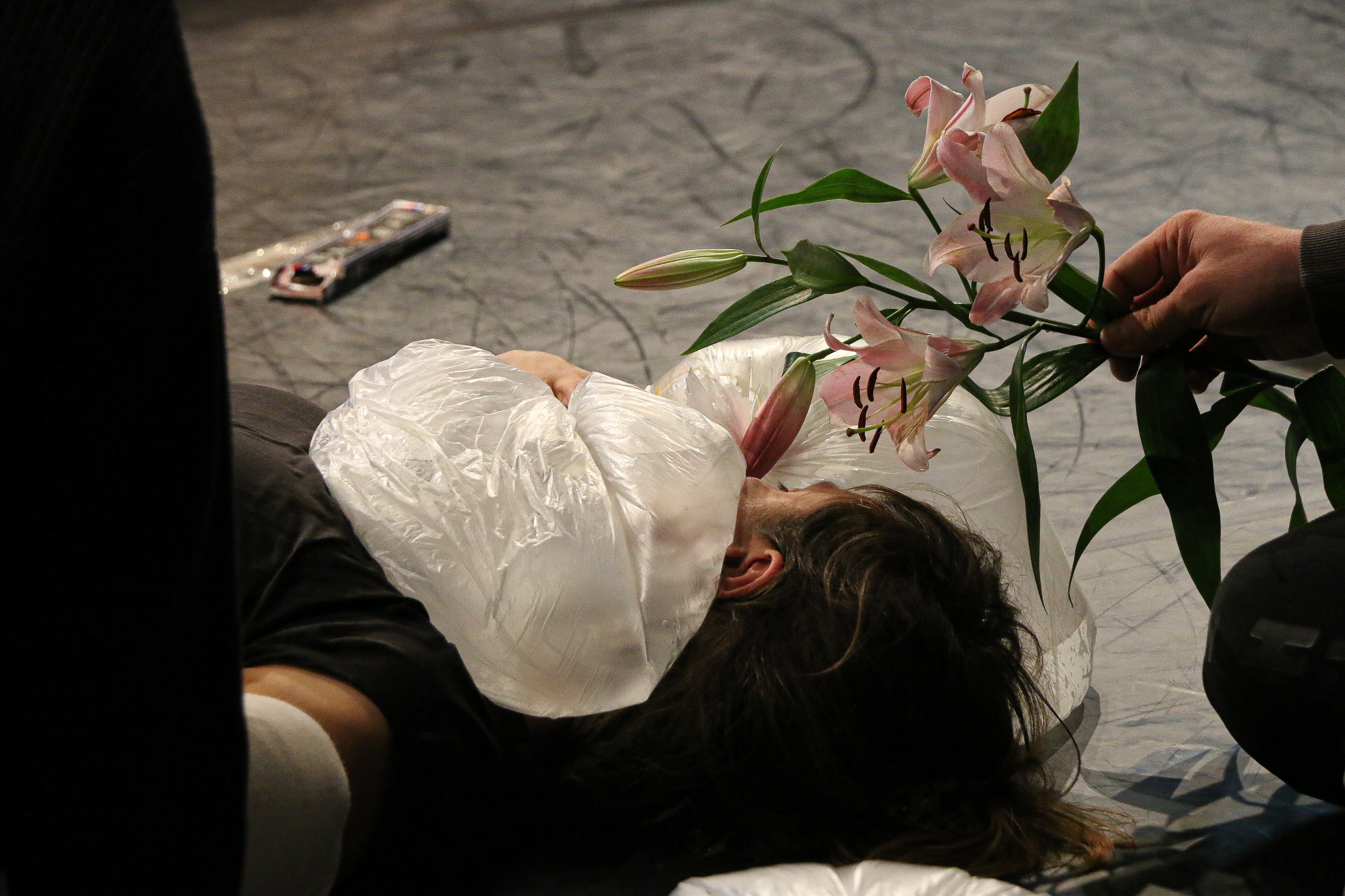 Eine Person liegt auf einem dunklen Boden. Auf ihrem Oberkörper liegen mit Luft gefüllte, durchsichtige Plastiksäcke. Eine andere Person hält ihr Blumen über das Gesicht.