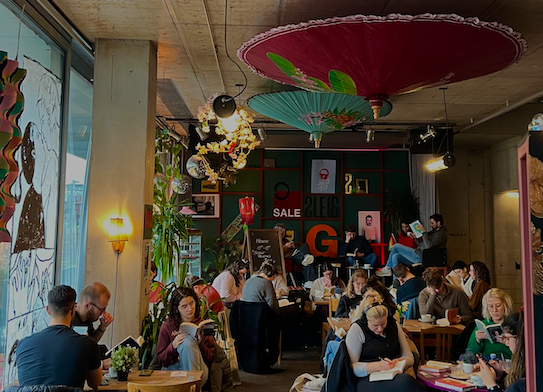 Man sieht den Innenraum eines gemütlichen Cafes. Vielen Menschen sitzen an Tischen und lesen.