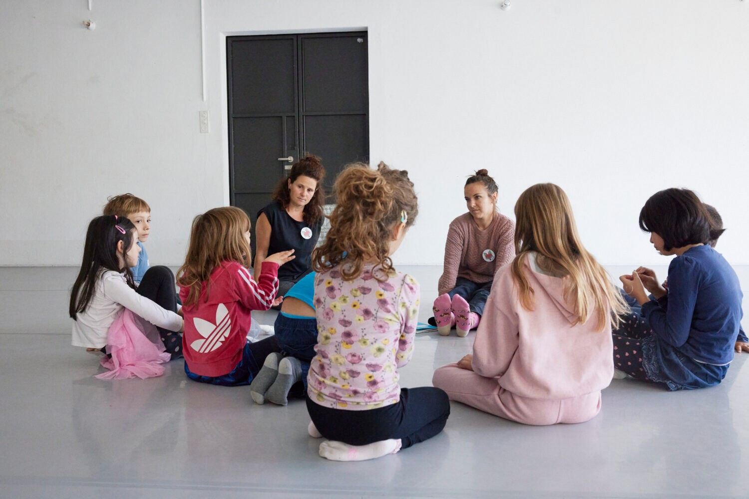 Man sieht einen modernen Raum. Auf dem Boden sitzen zwei Erwachsene und ein paar Kinder im Kreis. Ein Kind spricht, die anderen hören zu.