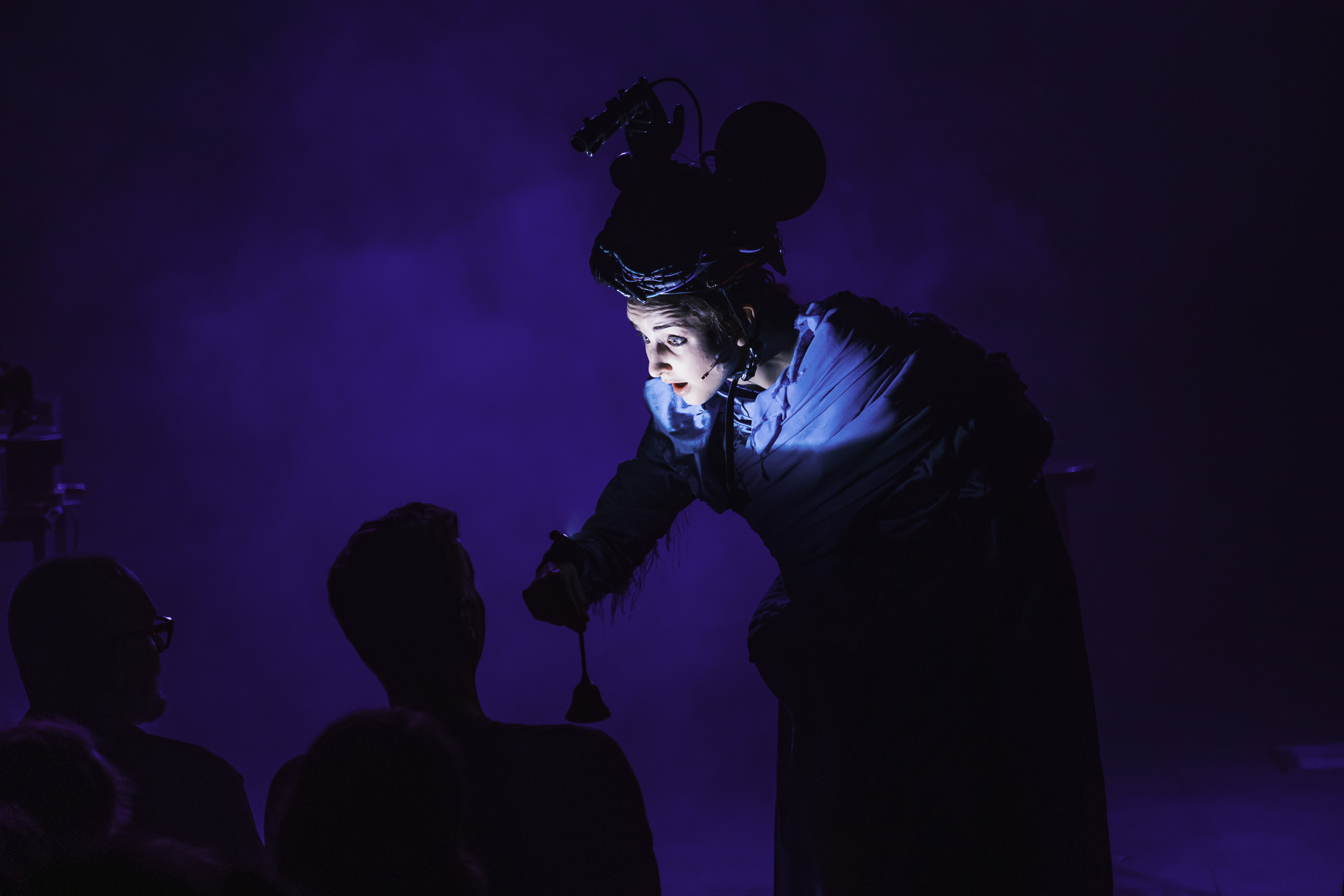 Im Dunkeln wird eine Person direkt angeleuchtet. Sie hat einen grossen Apparat auf dem Kopf und beugt sich zum Publikum.