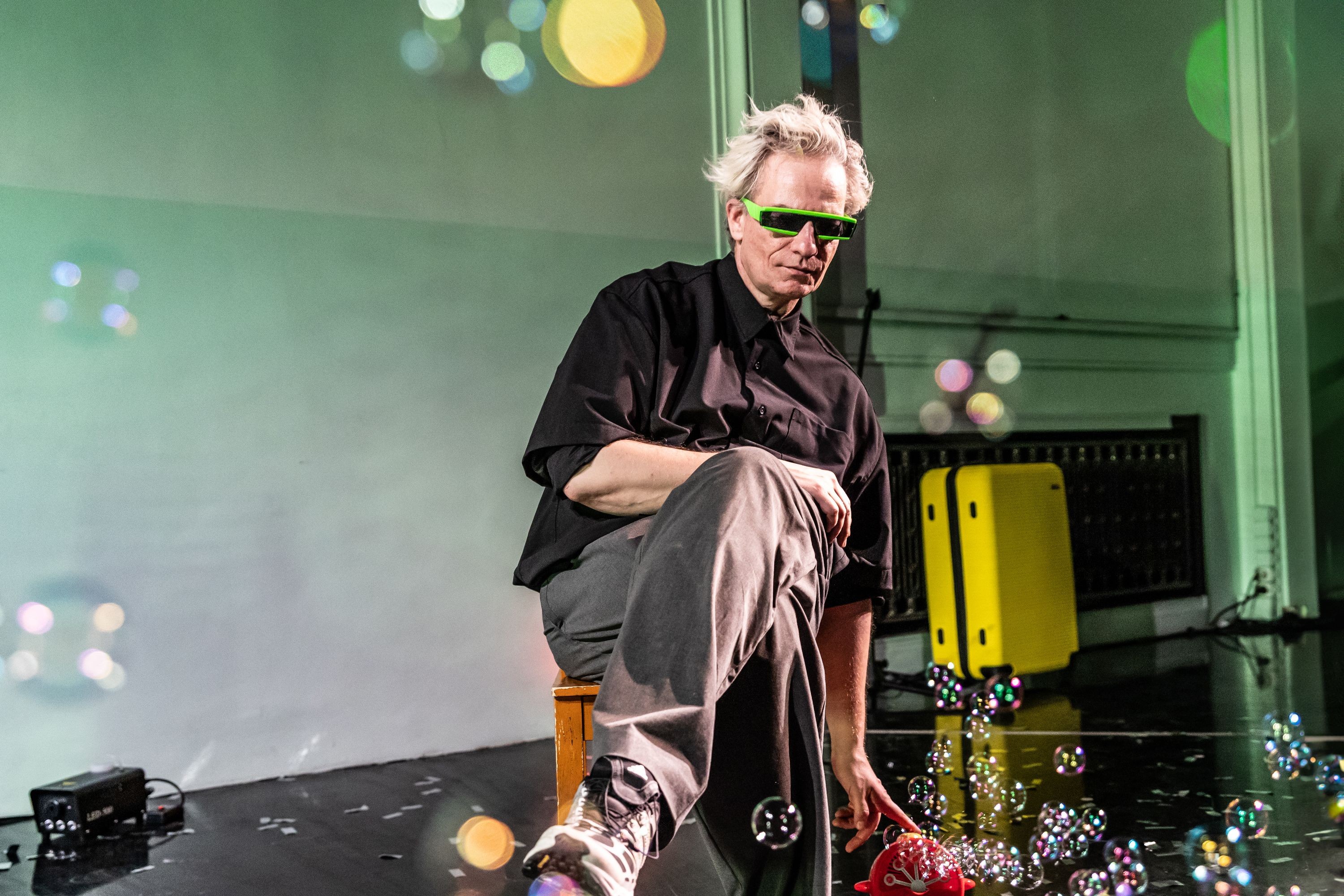 Der Performer sitzt auf einem Stuhl und drückt mit einer Hand auf eine Seifenblasenmaschine. Er trägt eine grüne schmale Sonnenbrille mit schwarzen Gläsern.