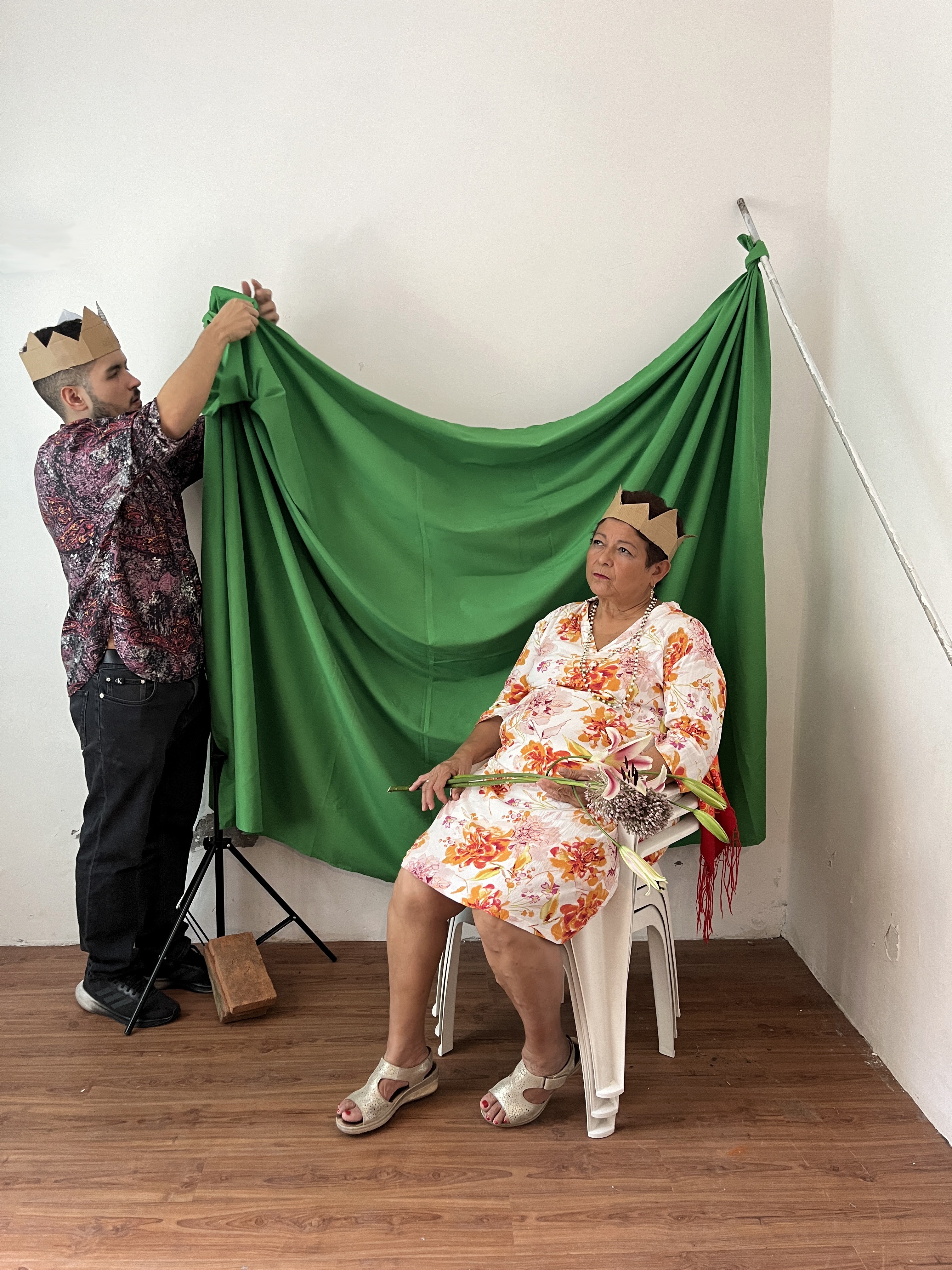 eine ältere Person sitzt auf drei übereinandergestapelten weissen Plastikstühlen. Im Hintergrund hängt eine jüngere Person ein grünes Tuch als Hintergrund auf. Beide tragen Kartonkronen