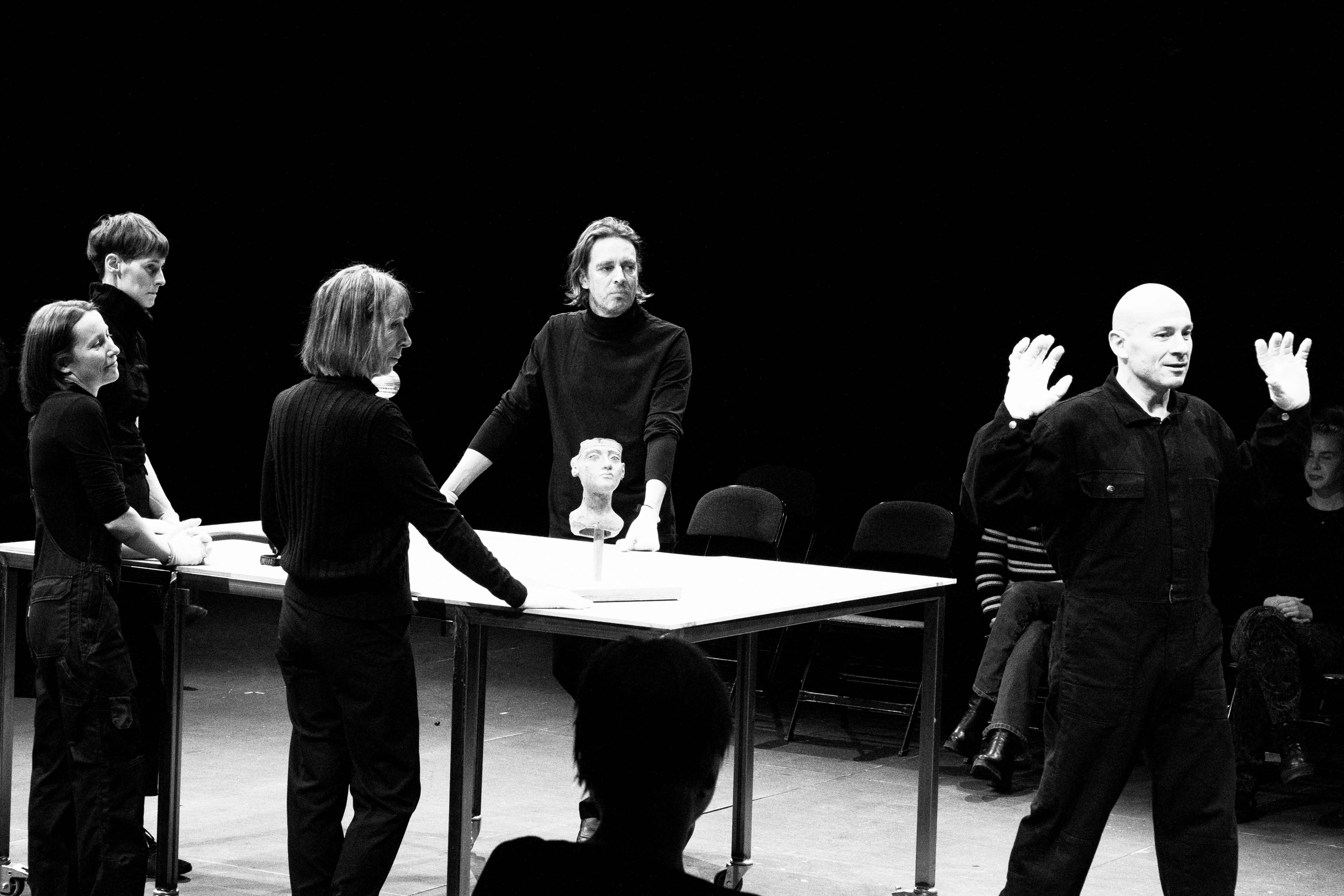 auf einem schwarz-weiss Bild stehen wir Personen in schwarzer simpler Kleidung um einen Tisch und schauen and das Ende. Dort hat eine fünfte Person den Rücken zu ihnen und hält die Hände hoch.