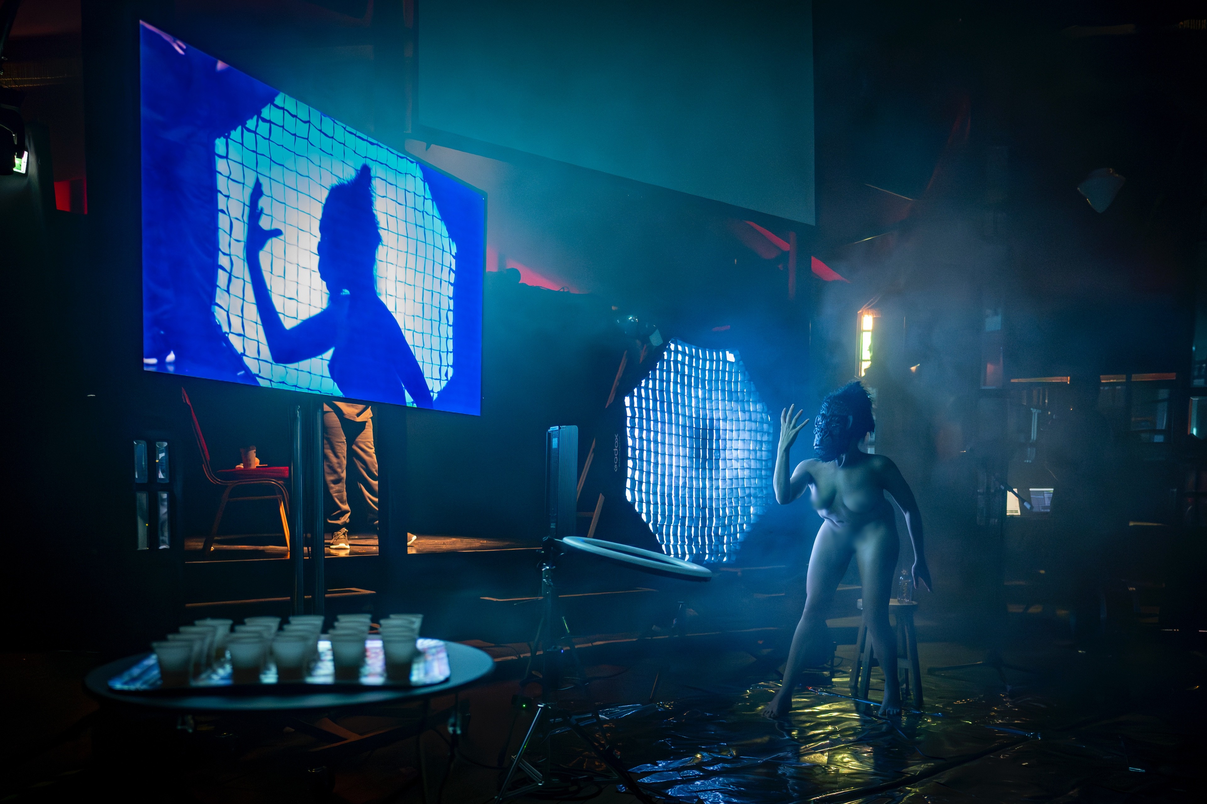 in einem unübersichtlich vollem Raum mit blauen Lichtern steht eine Person nackt und mit Gorilla Maske und hält die Hand vors Gesicht.