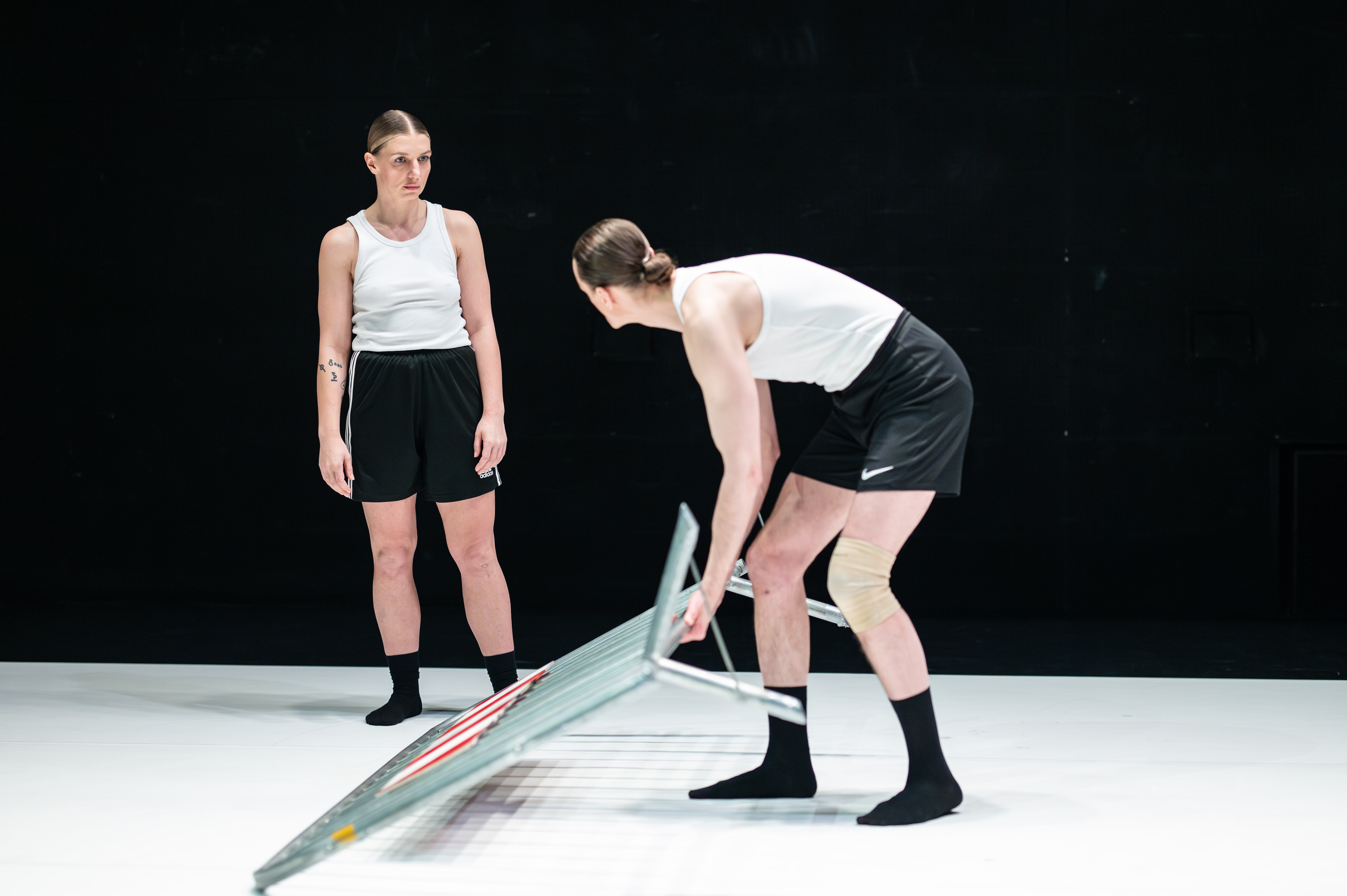 Die beiden Performer*innen stehen sich gegenüber. Der Performer hebt ein auf dem Boden liegendes Absperrgitter an und schaut der Performerin dabei in die Augen.
