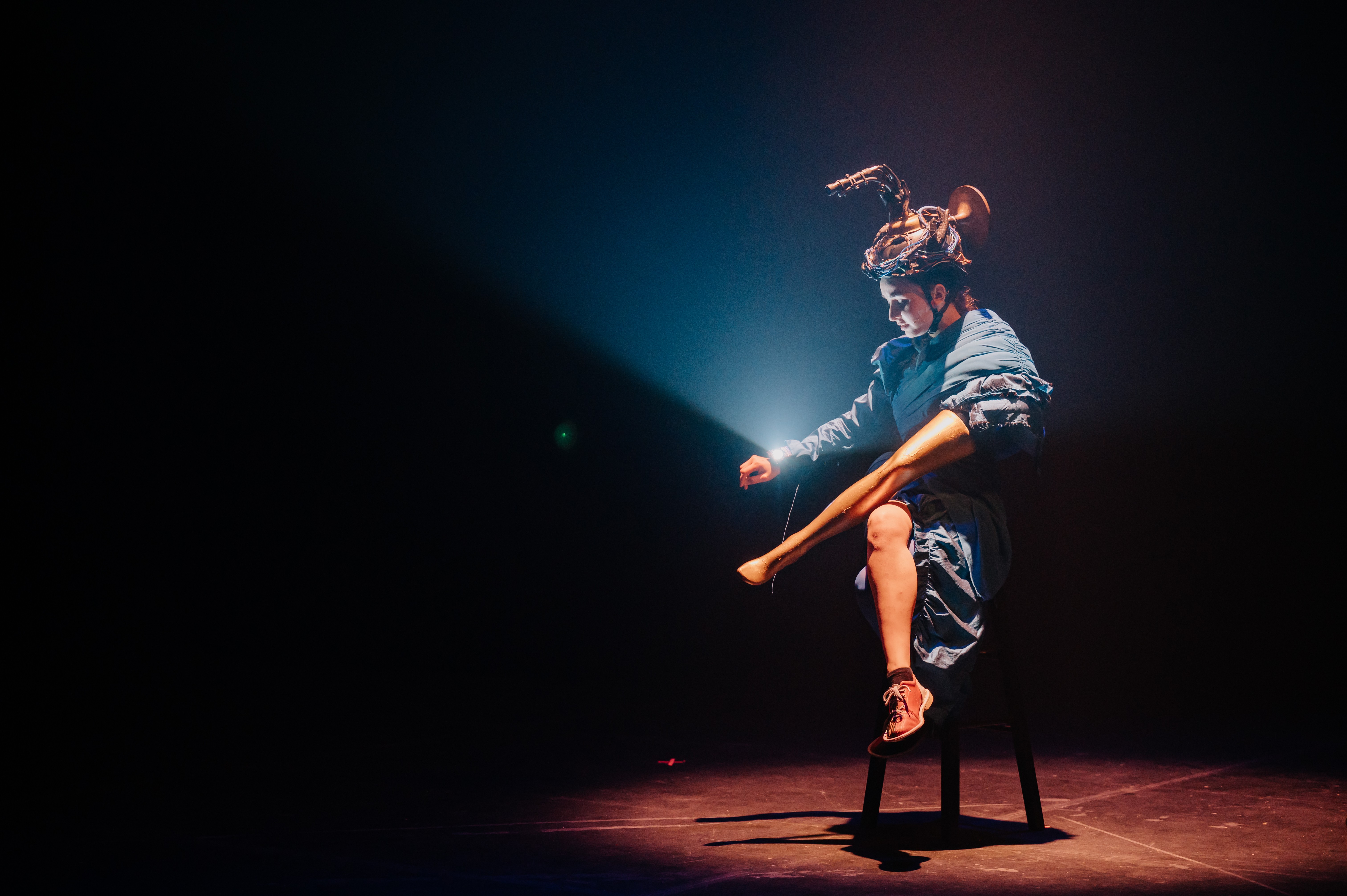 eine Person sitzt in schumrigem Licht auf einem Hocker. Ein Arm hat sie in einem Puppenbein drin und verkreuzt damit ihre Beine. Auf ihrem Kopf hat sie einen Apparat und sie hat eine Uhr an, die Licht macht.