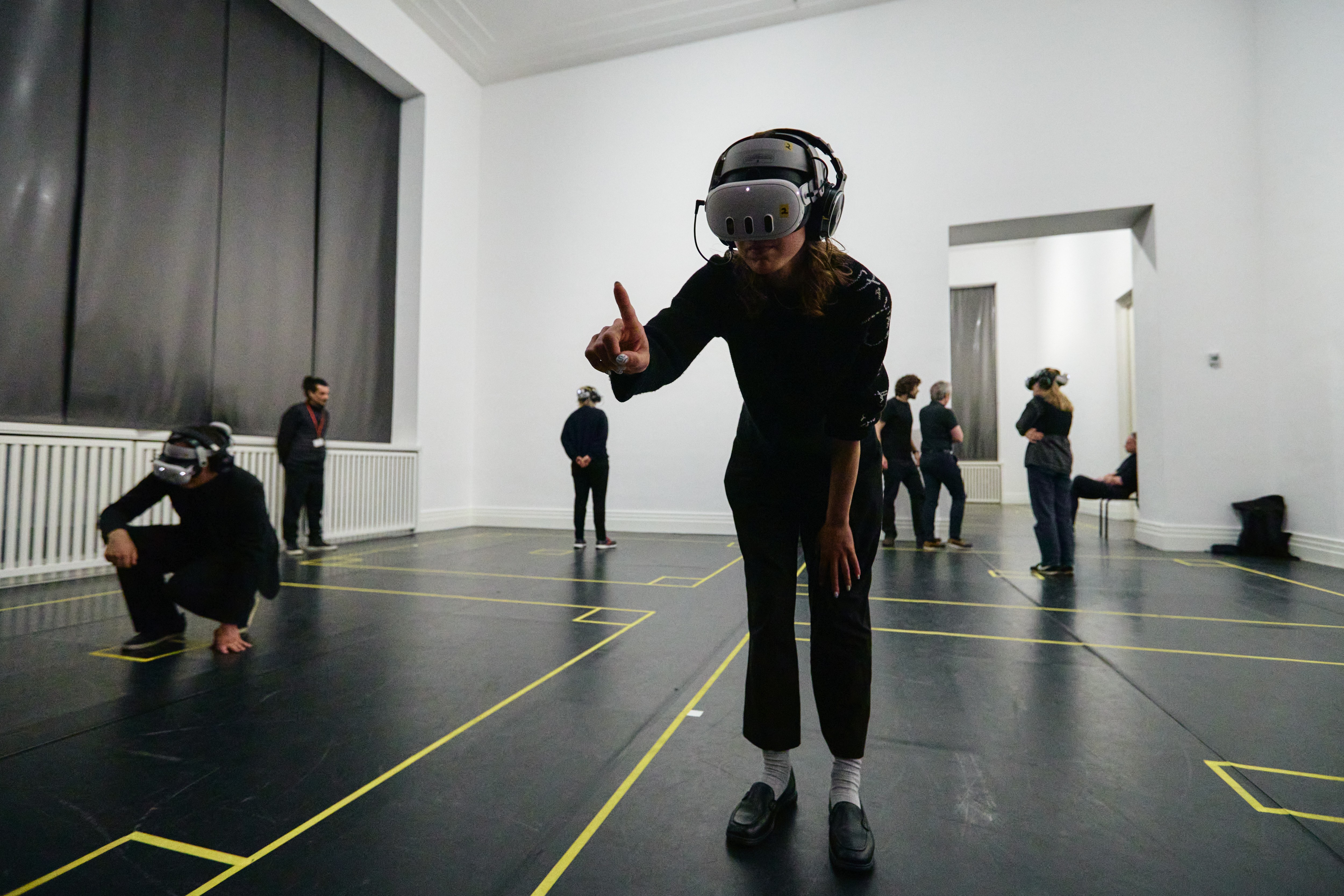 Ein Raum mit schwarzem Boden und Markierungen die es in Boxen aufteilt. Mehrere Personen haben VR Brillen an und interagieren mit dem Spiel