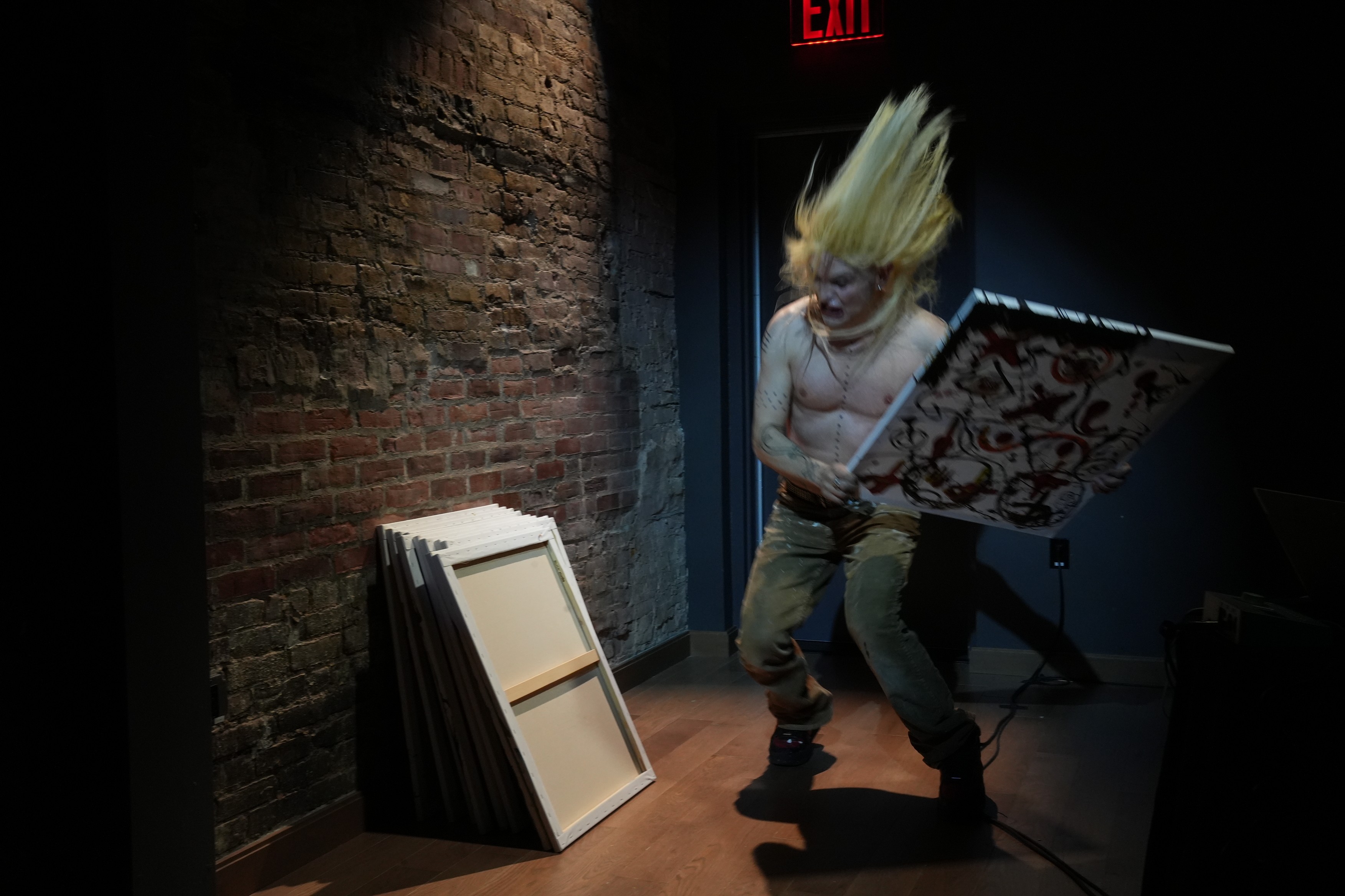 eine leichtbekleidete Person hält ein schwarz-weisses Gemälde in den Händen und ist in Bewegung, es an die Wand zu werfen, wo mehrere Leinwände angelehnt sind.