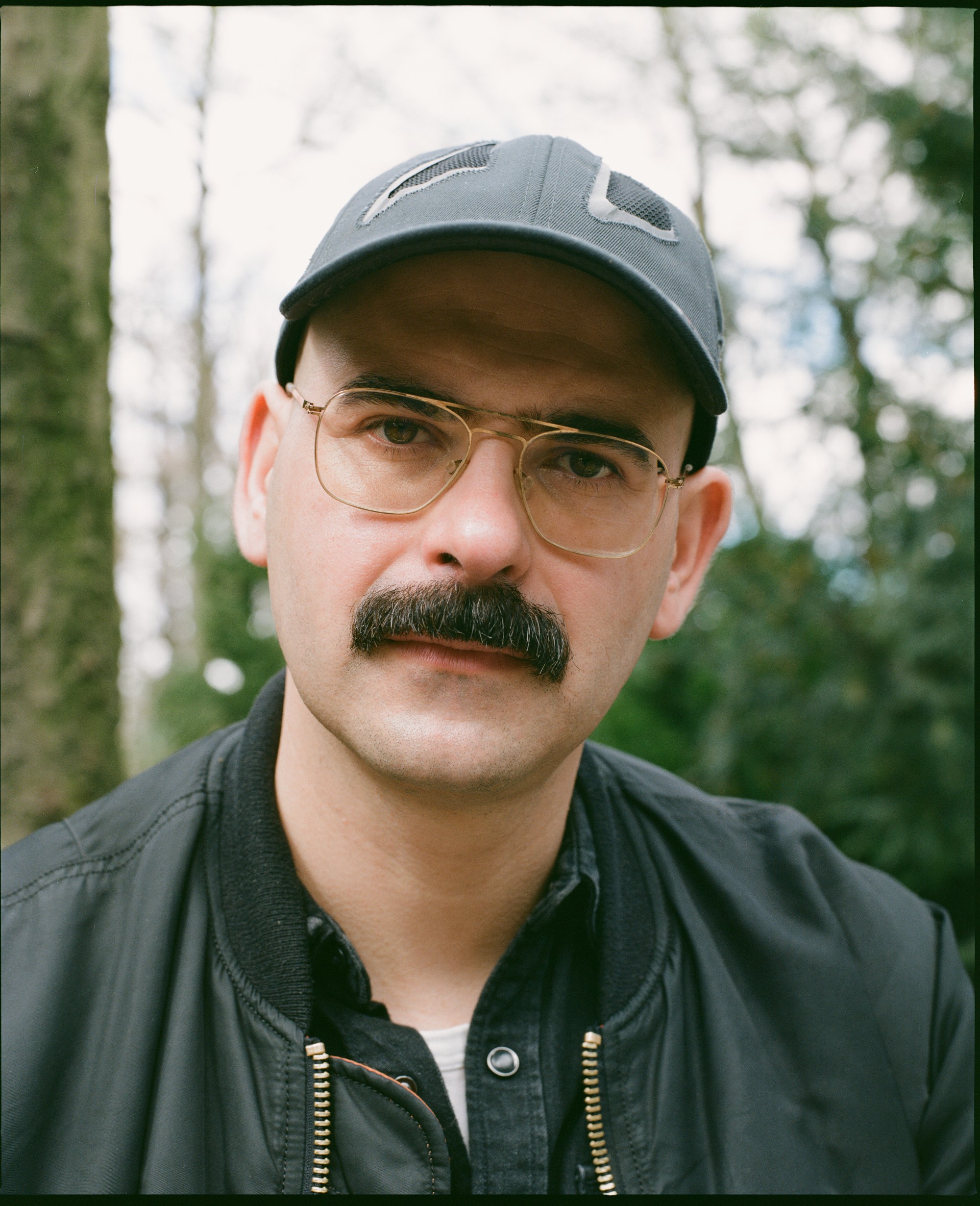 Ein Portrait von einer Person. Sie schaut neugierig in die Kamera, trägt schwarze Kleidung, eine Brille und hat einen Schnurrbart. der Hintergrund ist ein Wald