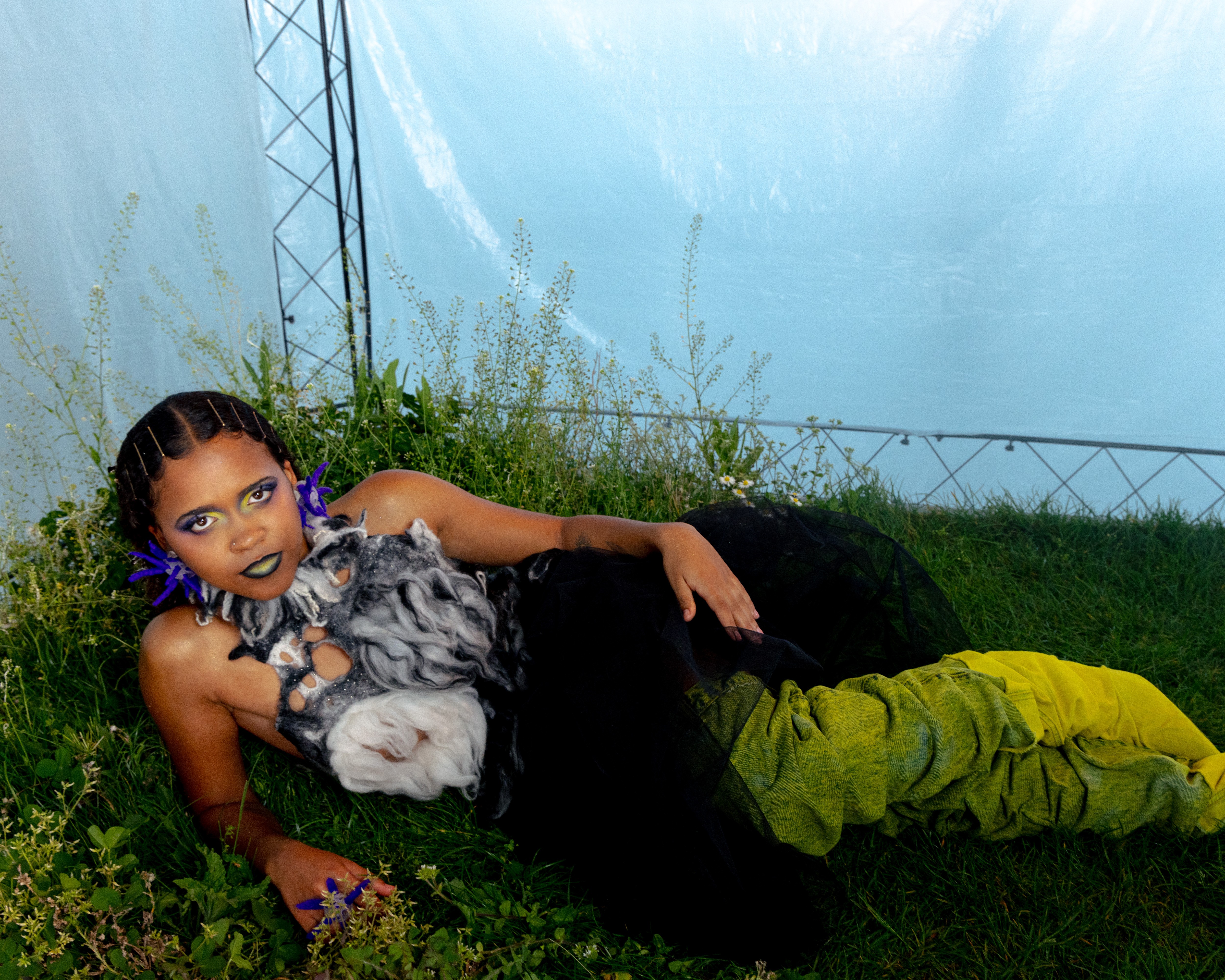 eine Person liegt vor einem Zelt-ähnlichen Hintergrund auf Gras. Sie ist elegant angezogen und schaut mit starkem Blick in die Kamera.