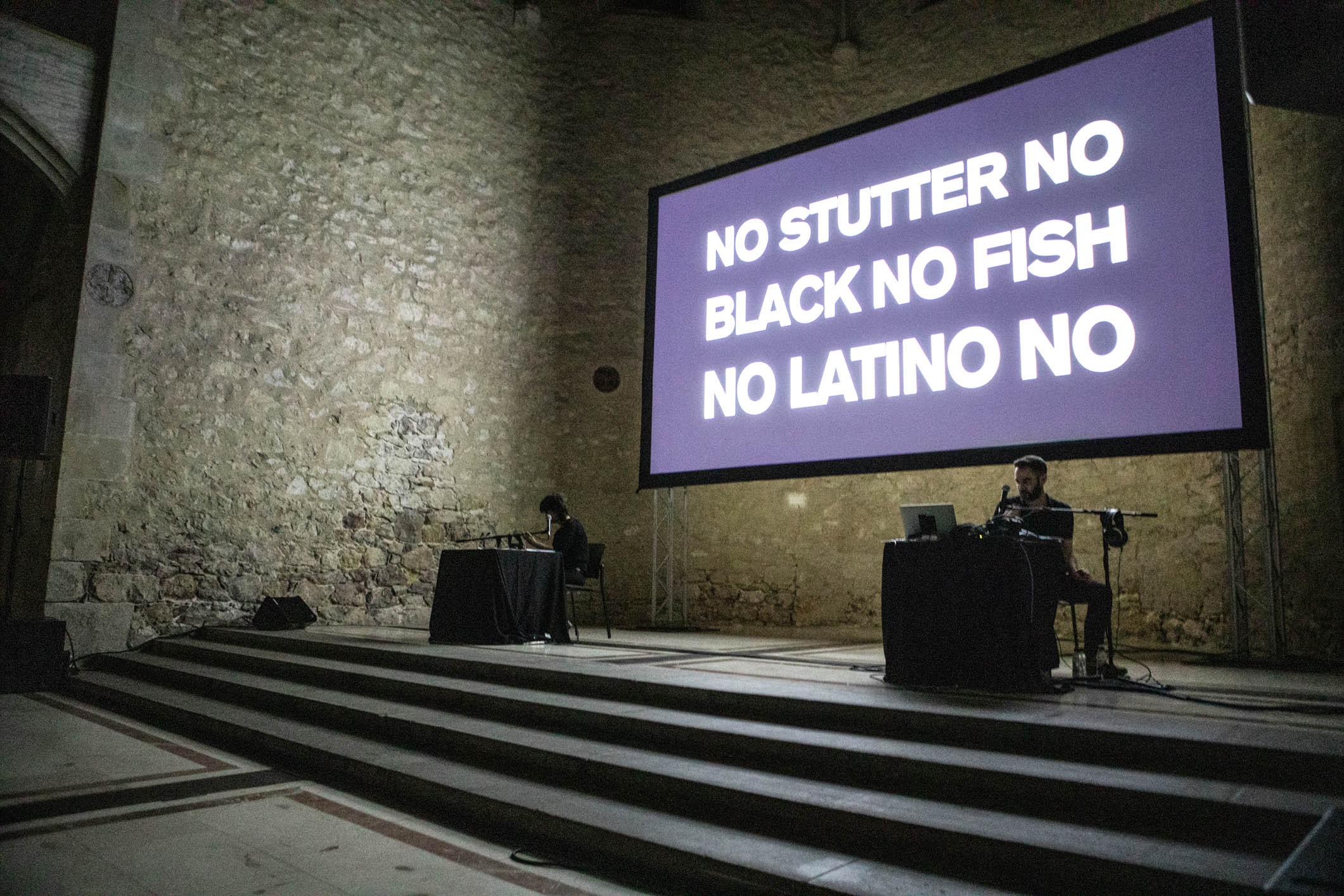 Der Raum ist gross und die Wände aus grauem Gestein. An einer dieser Wände ist eine grosse Leinwand mit der Aufschrift "No stutter no black fish no latino no». Vor der Leinwand sitzen zwei Personen je an einem Tisch mit Mikrofon.