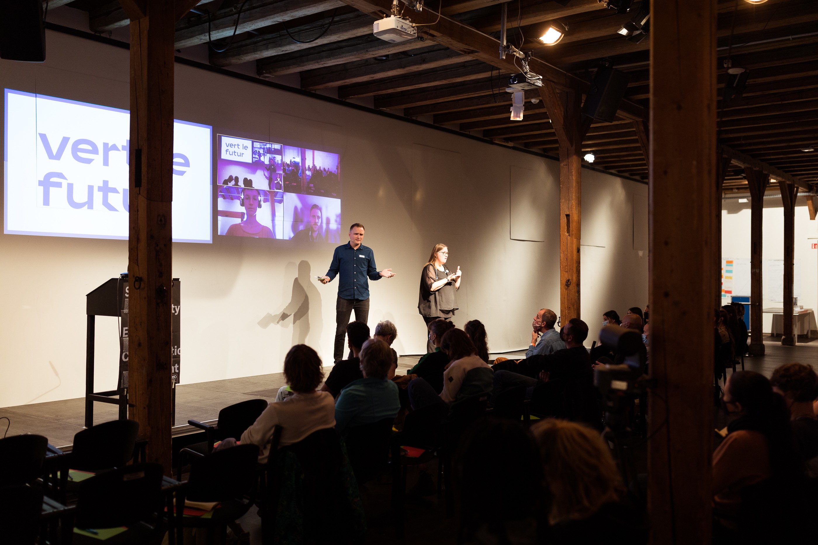 Das Bild zeigt einen abgedunkelten Raum mit Holzpfeilern. Ein kleines Publikum schaut auf eine Bühne, wo jemand einen Vortrag hält. An der Wand dahinter gibt es Projektionen.