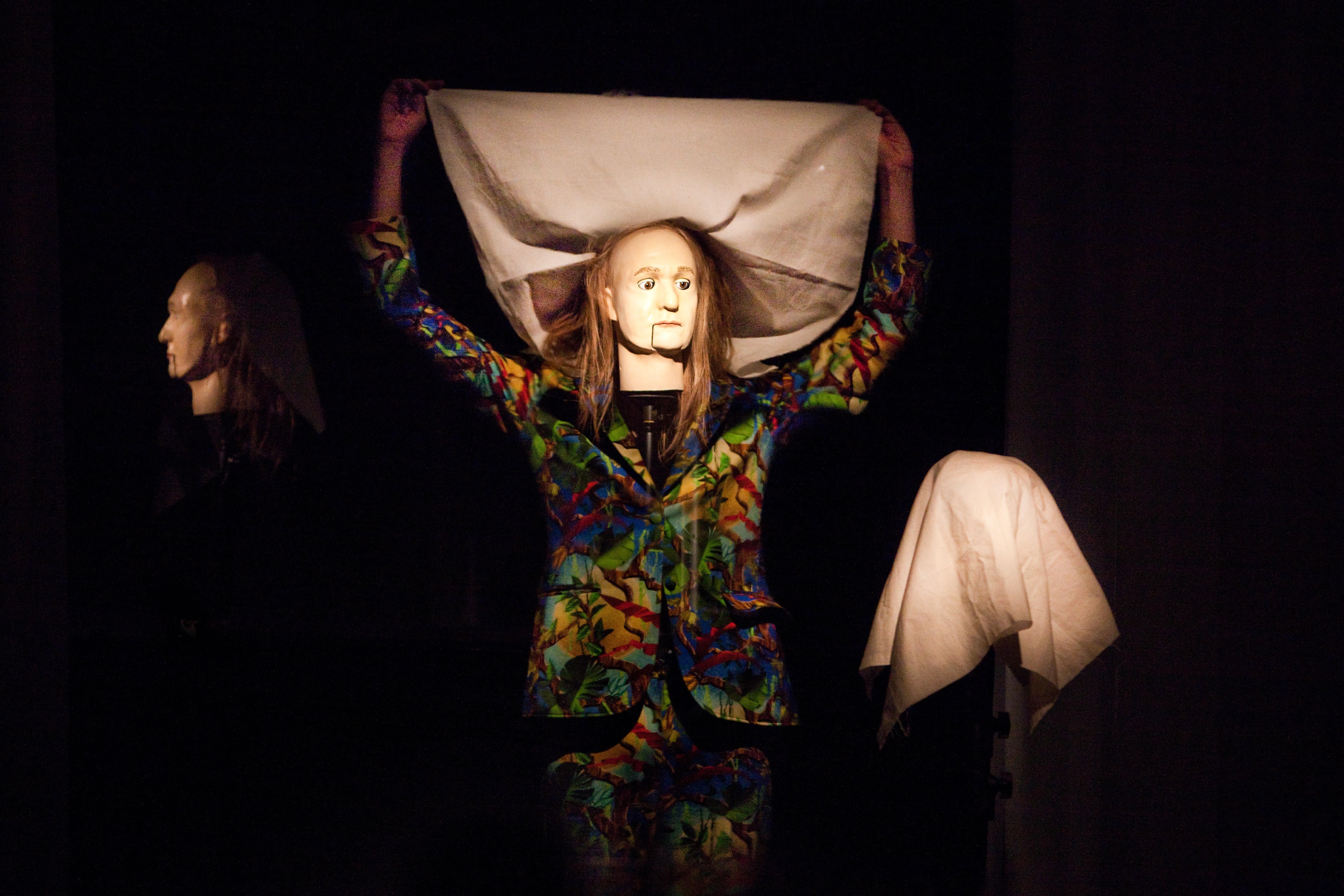 In der Mitte steht Diederik Peeters in einem sehr bunten Anzug. Diederik trägt eine Maske mit langen Haaren. Mit den Händen hält sich Diederik ein weisses Tuch wie ein Dach über den Kopf. Vor Diederik hängt ein weisses Tuch über einer Stange.