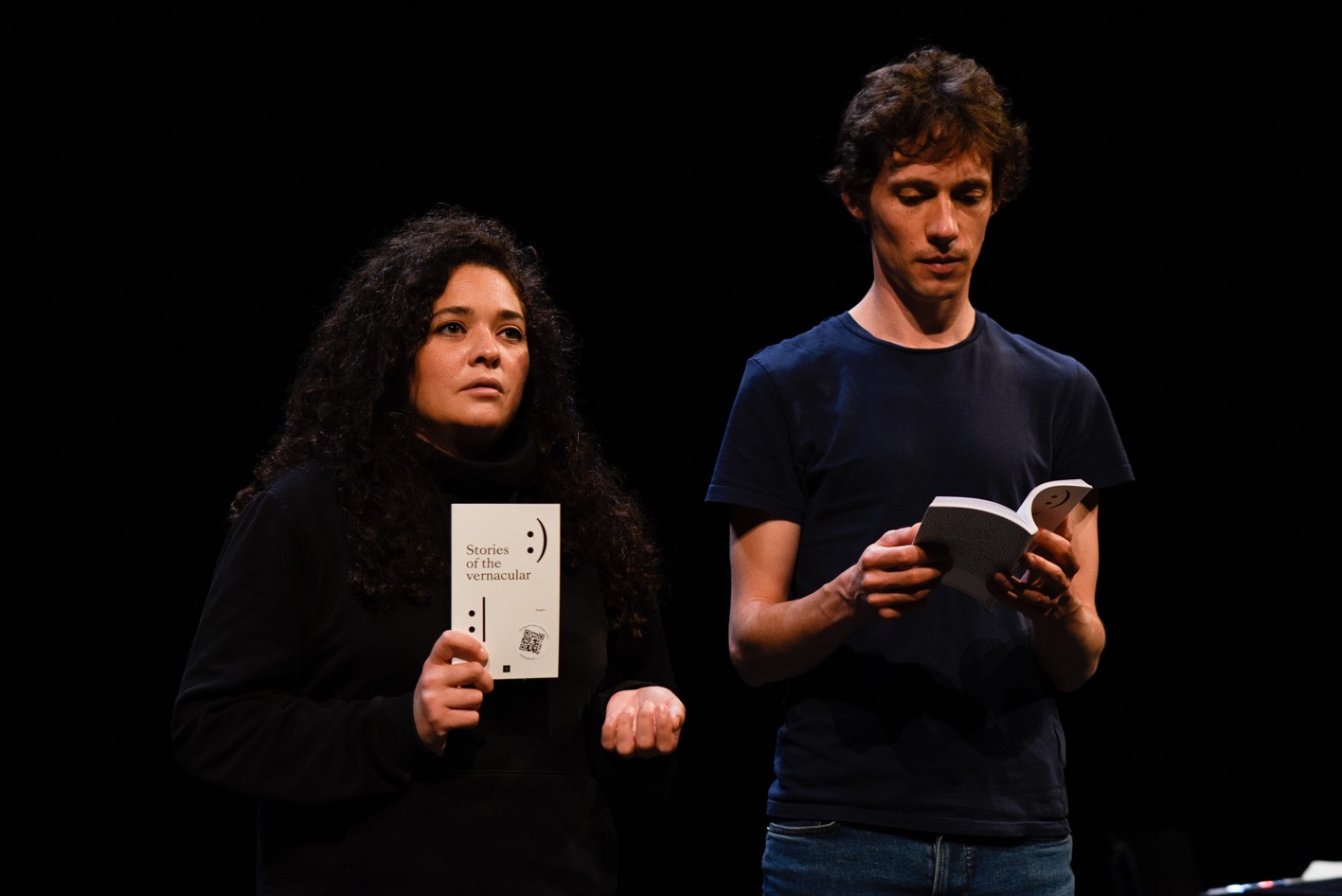 Der Hintergrund ist schwarz. im Vordergrund stehen zwei Personen. Die linke hält ein Papier mit der Aufschrift «Stories of the vernacular». Die rechte hält ein Buch in der Hand.