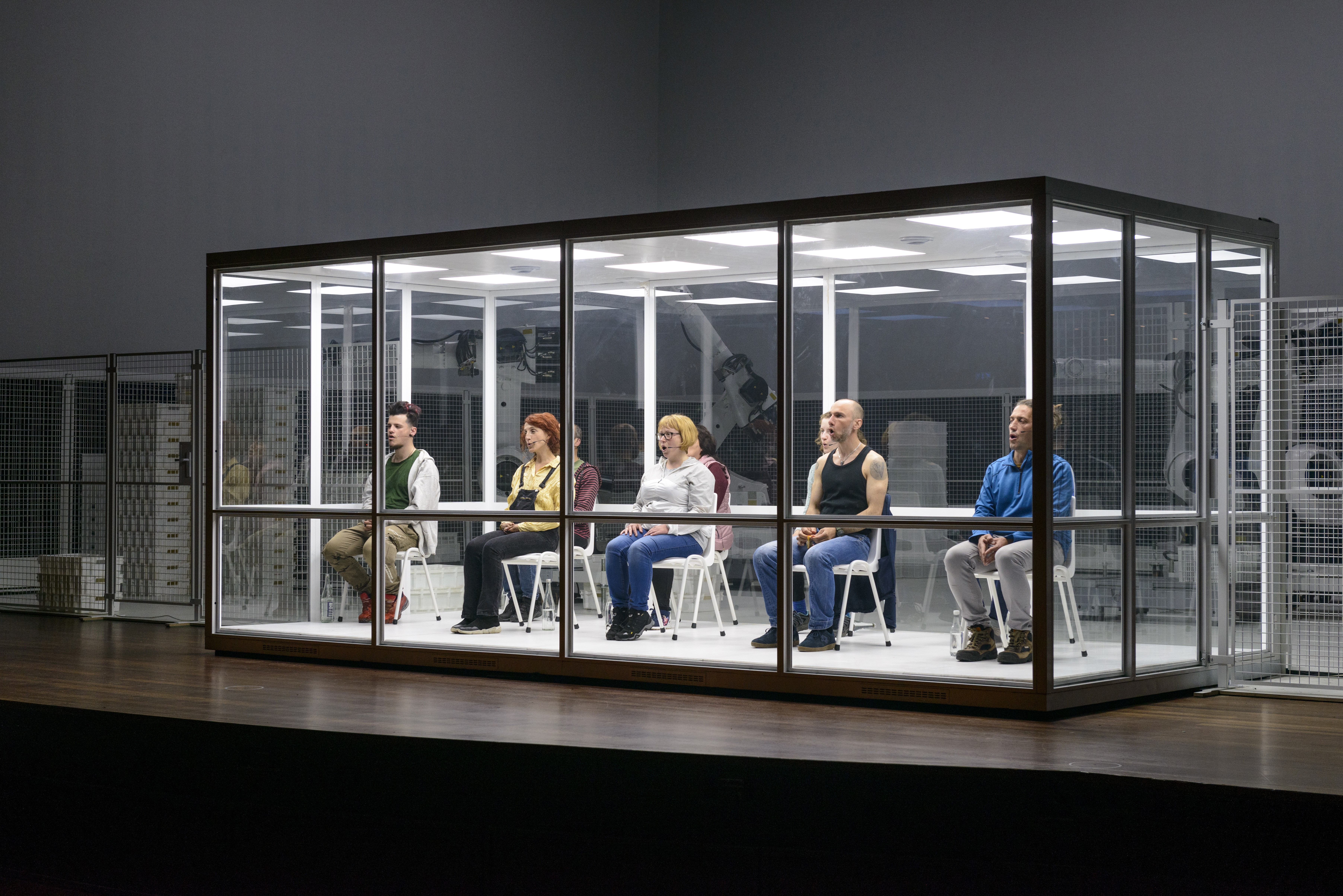Im Hintergrund weisse Wände und grosse weisse Roboter. Im Vordergrund eine grosse rechteckige Glasbox, in der 9 Personen auf weissen Stühlen sitzen und singen.