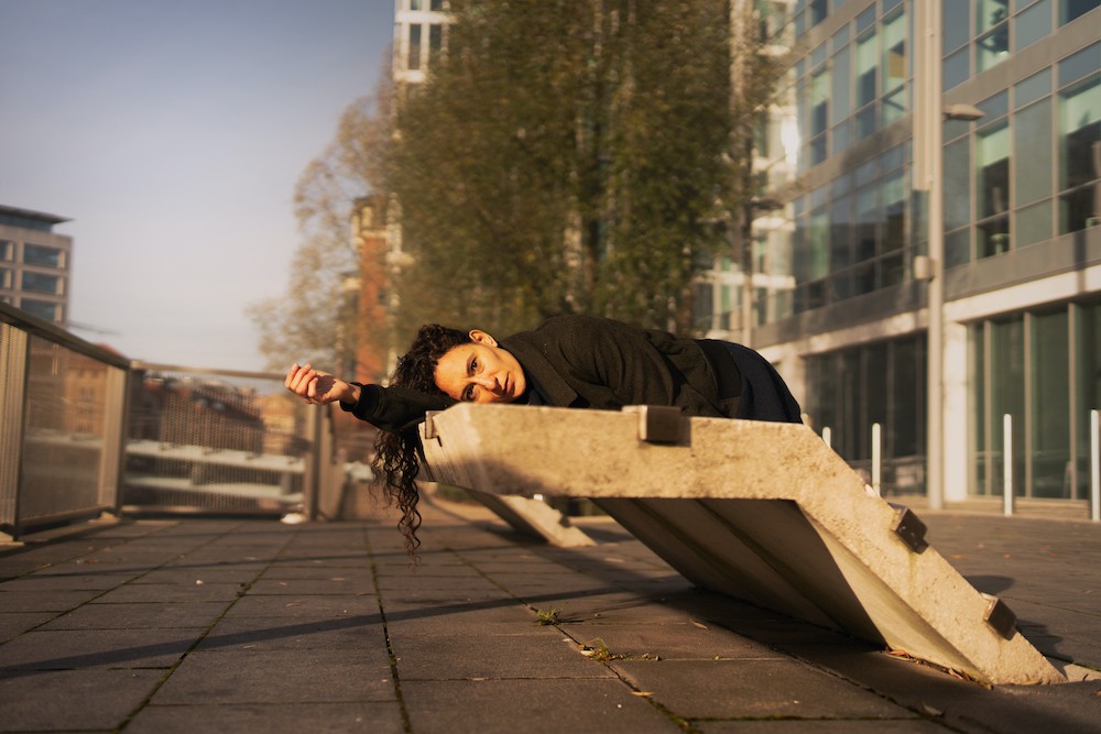 Im Hintergrund moderne Gebäude und Bäume. Im Vordergrund liegt eine Person mit langen Haaren auf einem Betongebilde, die Sonne scheint ihr ins Gesicht.