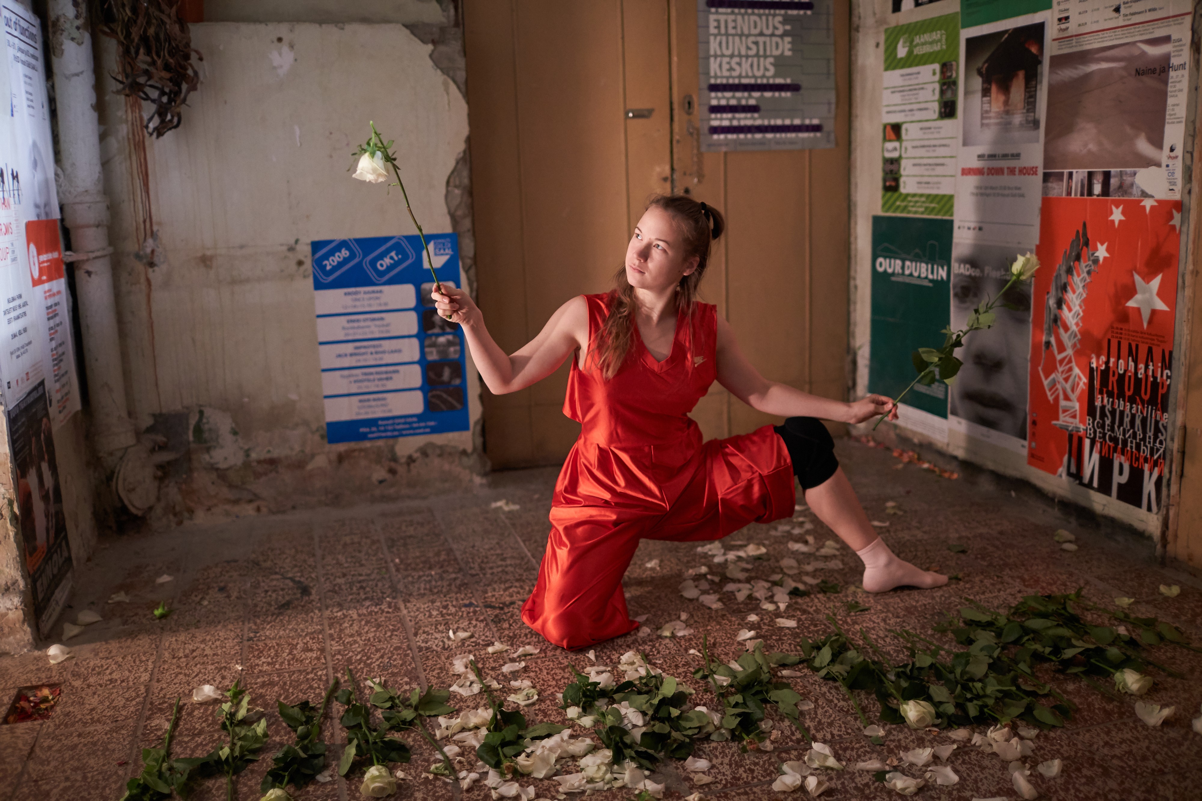 Ein Raum, der mit verschiedenen Plakaten ausgekleidet ist. In der Mitte des Raumes eine Person in rotem Kleid, die weisse Rosen hält. Vor ihr hat es viele weisse Rosenblüten.