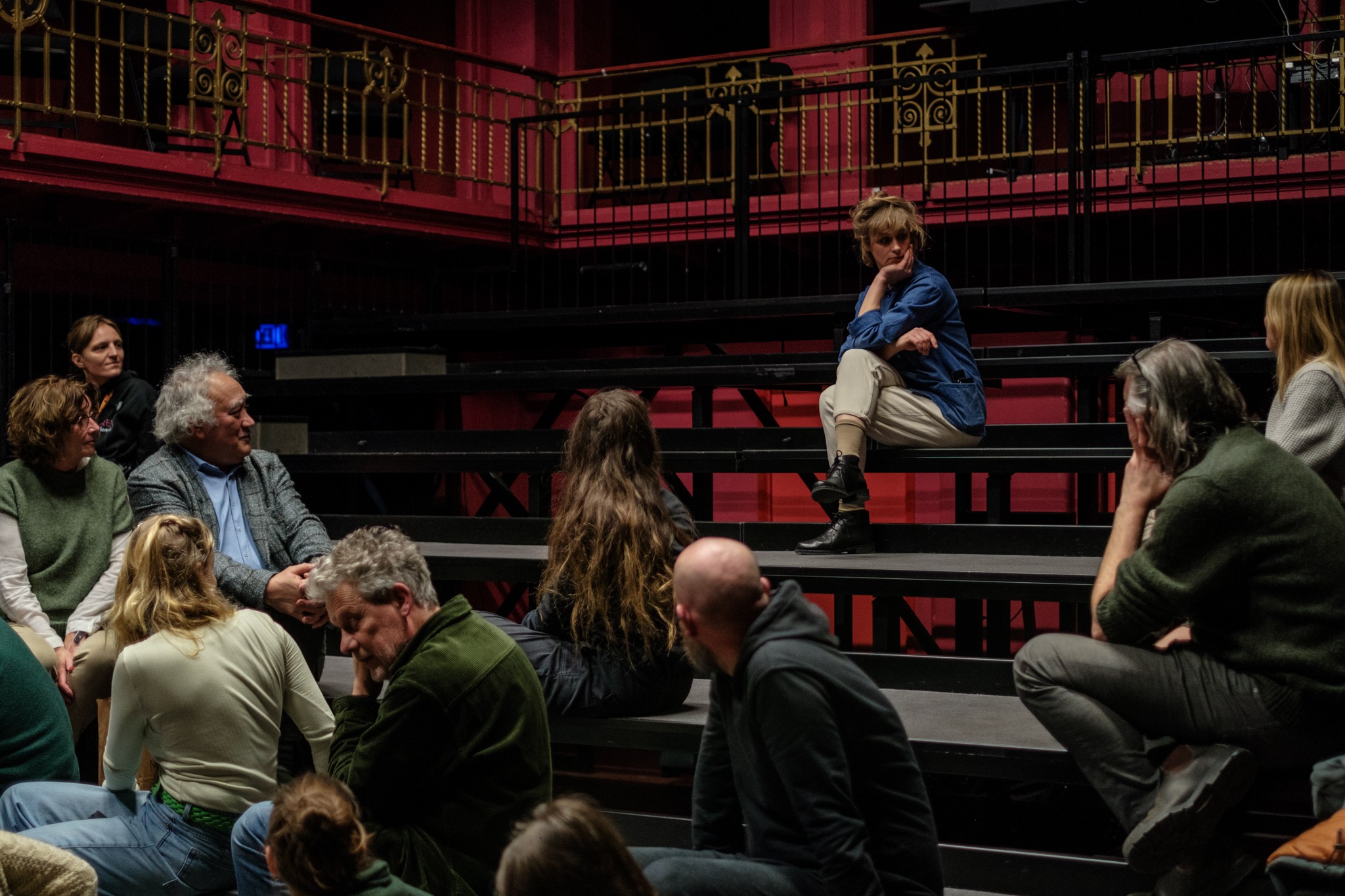 Man sieht eine Theatertribüne. Eine Person sitzt allein auf einer Bank und schaut die anderen Menschen an.