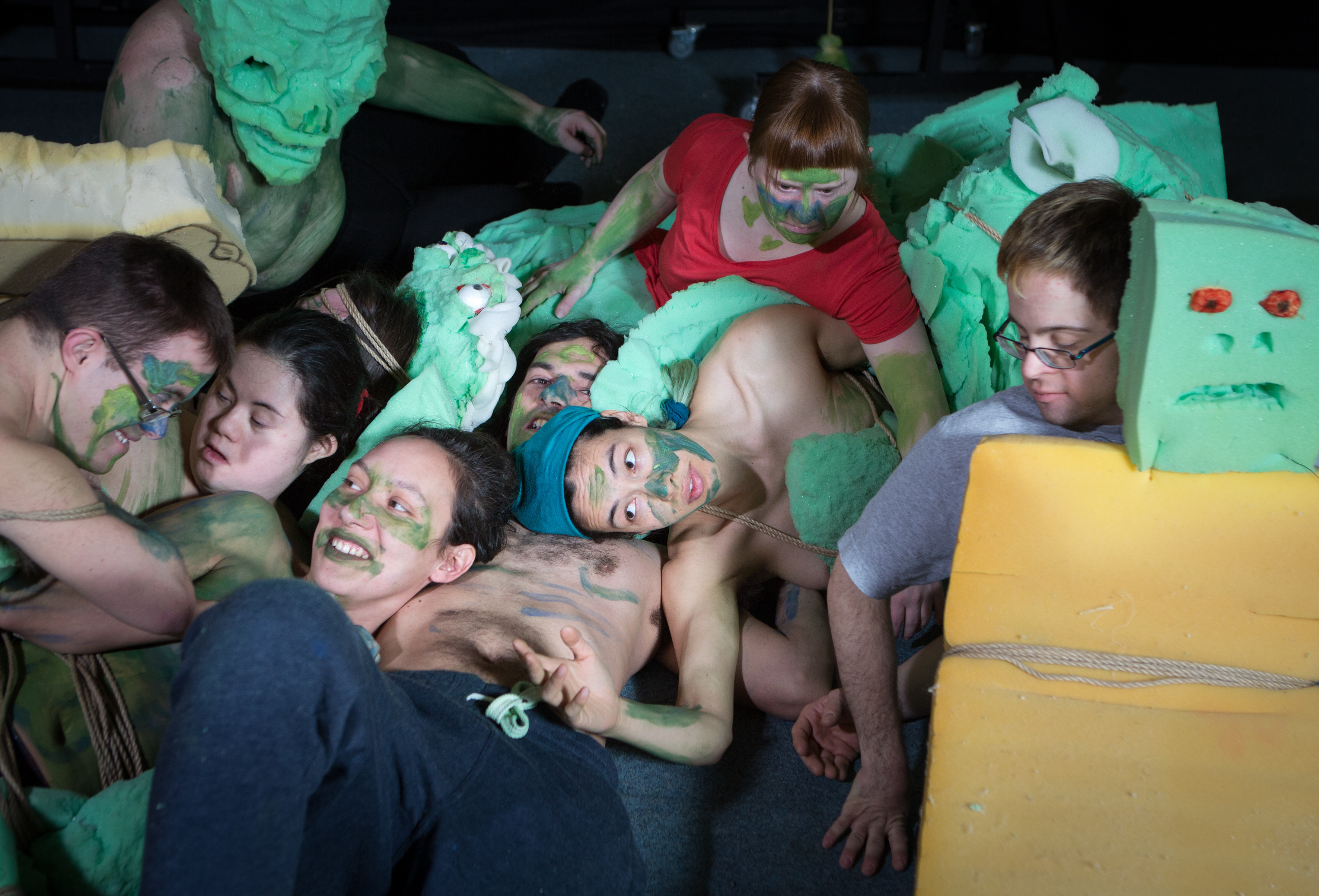 Sieben Menschen liegen in einer Gruppe, sie haben die Gesichter teilweise grün angemalt, auf dem Boden liegt grüner Schaumstoff