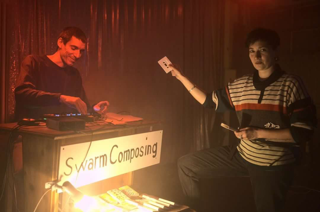 Das Bild zeigt die DJs mit Namen Plouk, die an einem Mischpult in einem dunklen Raum stehen. Eine Person hält eine Kassette in die Luft. Auf einem grossen Schild steht "swarm composing".