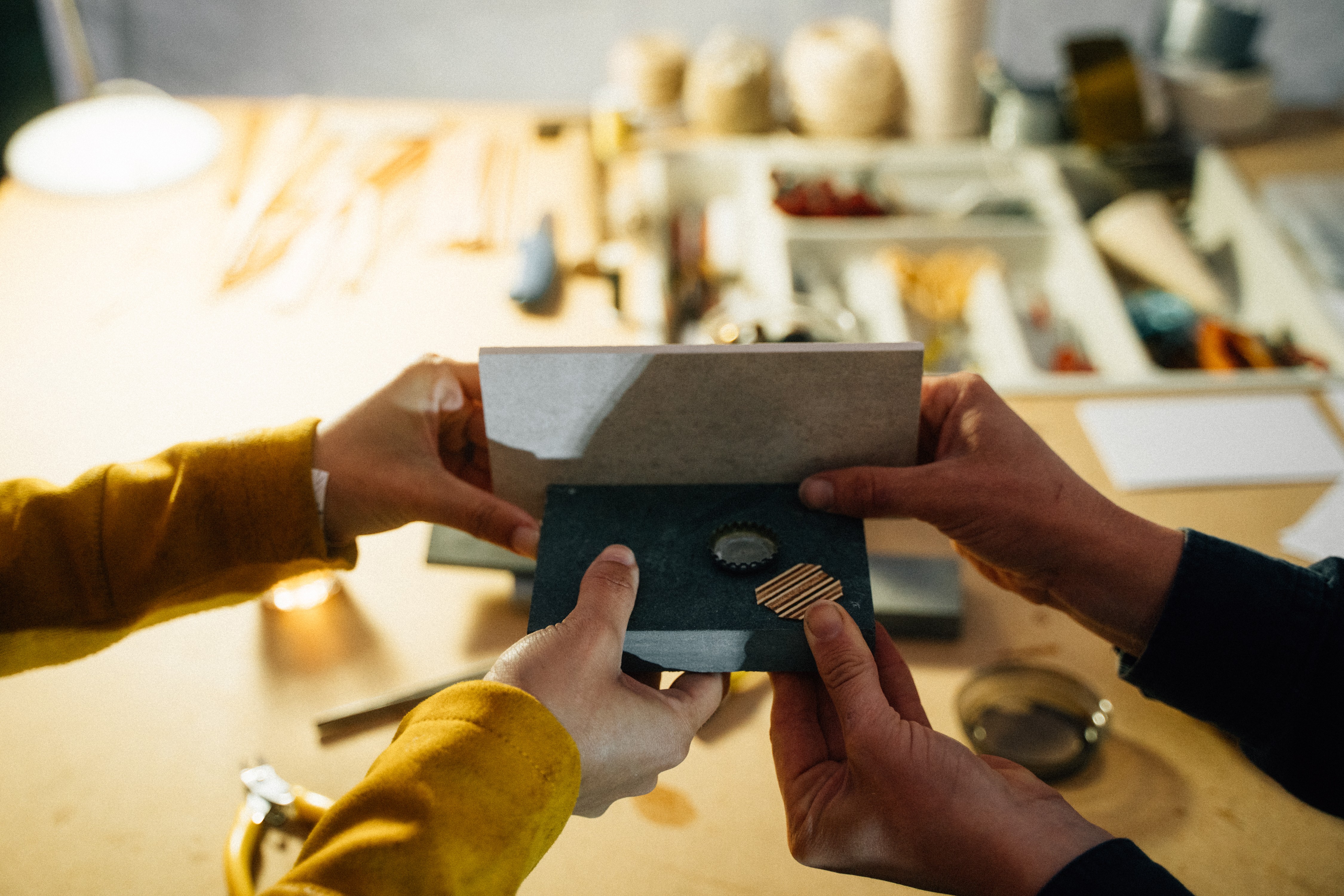 Im verschwommenen Hintergrund sieht man einen Tisch mit verschiedenen Materialien und Werkzeugen. Im Vordergrund sieht man vier Hände, die einen abstrakten grauen Gegenstand halten.