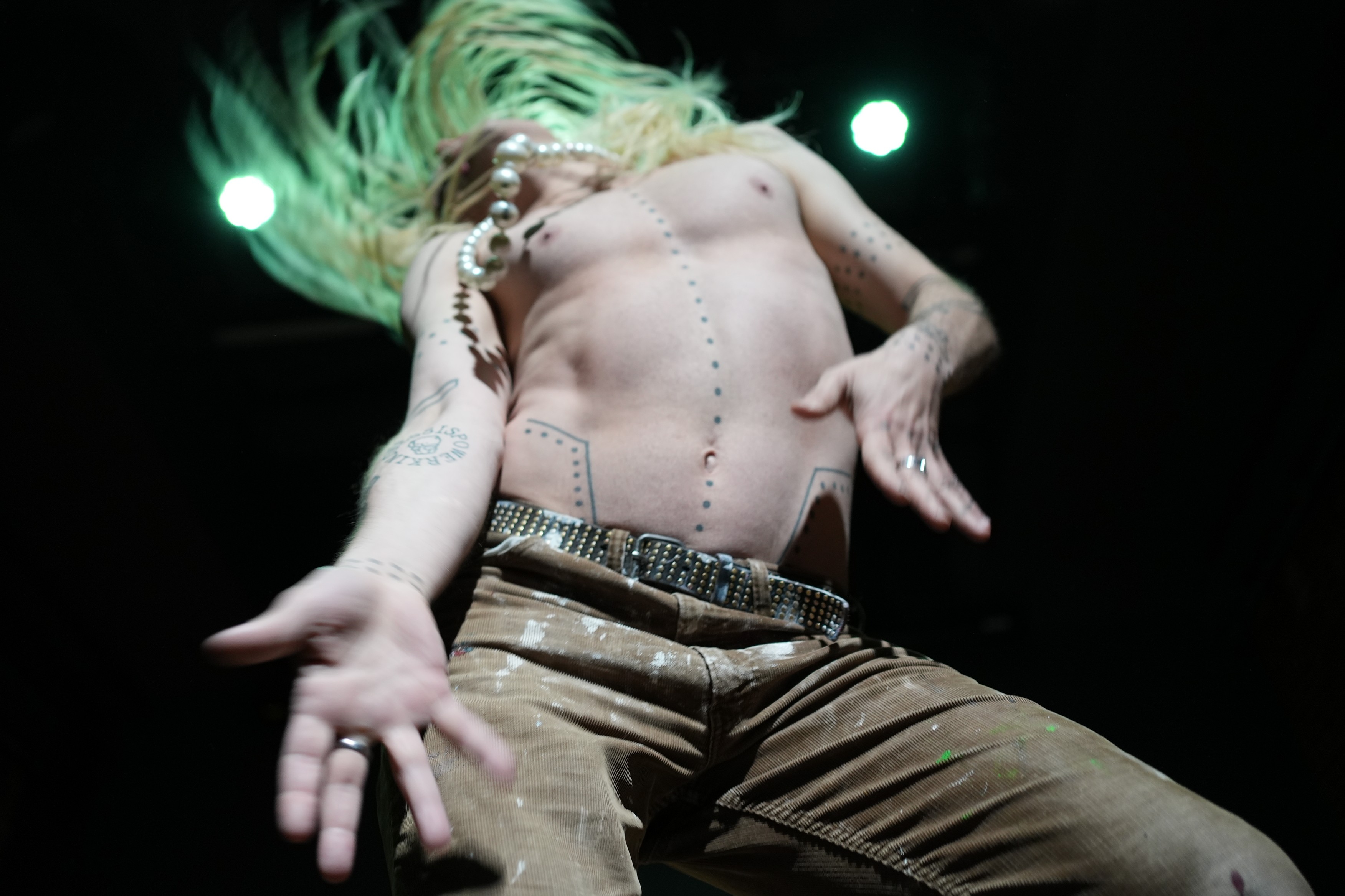 eine leichtgekleidete Person schwingt lange vom Licht grüne Haare und tanzt. Das Gesicht ist weggedreht.