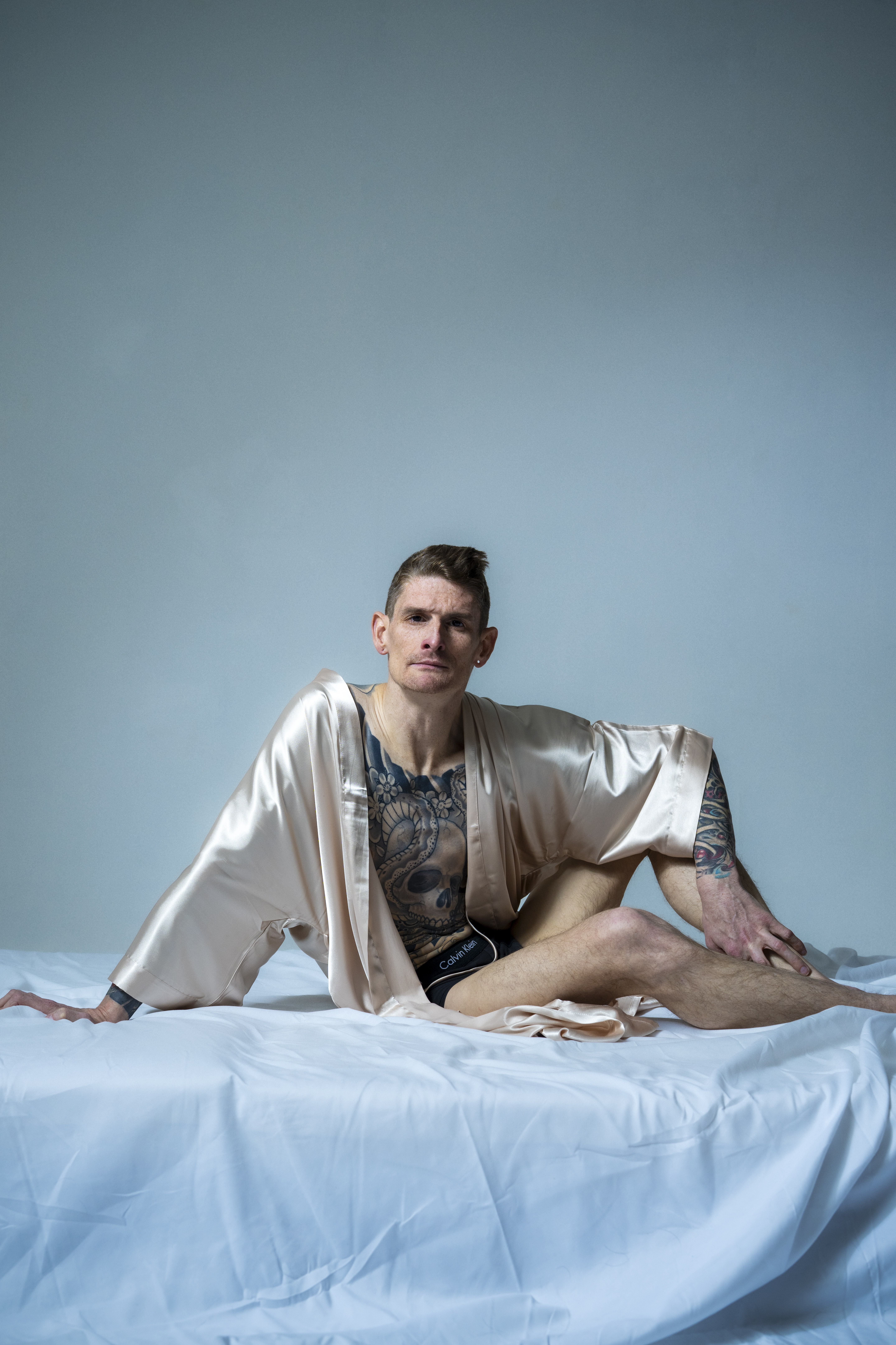 Ein Performer sitzt auf eine weissen Lacken mit nur einem goldenen Bademantel bekleidet und schaut ernst in die Kamera.