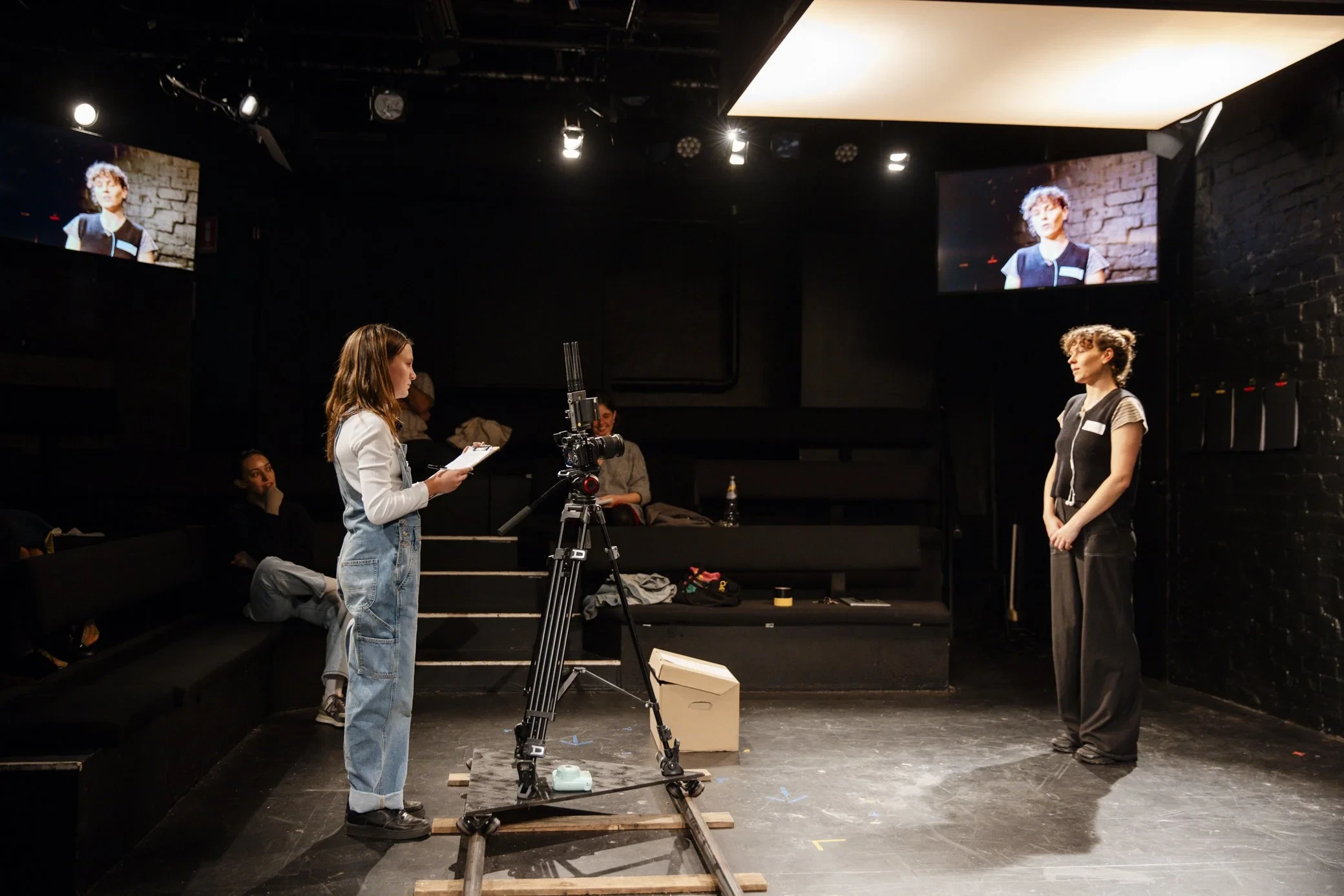links auf der Bühne steht ein Kind mit einem Block in der hand neben einem Kamera Set-Up. Dieses filmt eine andere Person, die direkt in die Kamera spricht. Das Video wird auf einem Bildschirm hinter der interviewten Person projiziert.