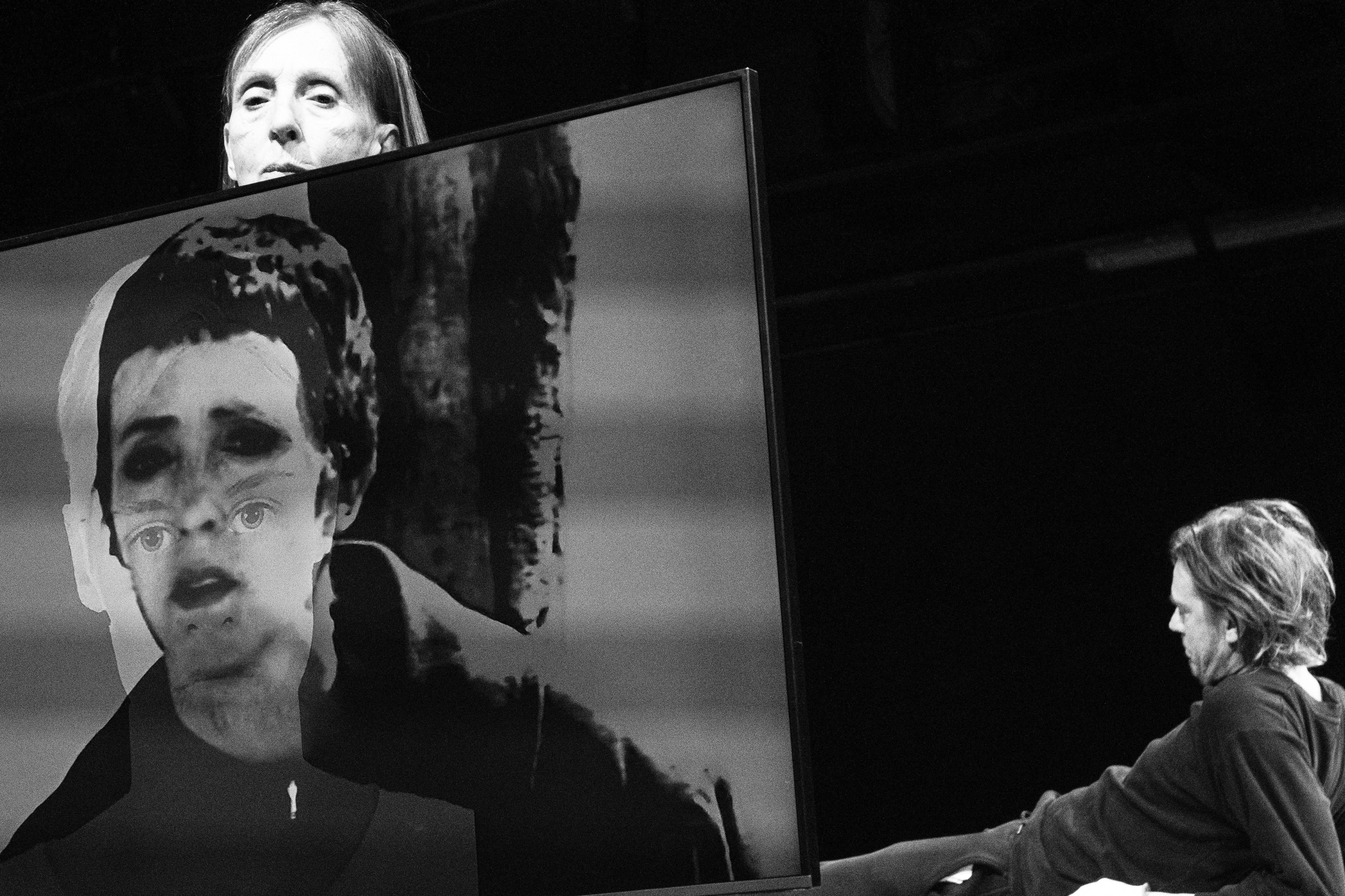 auf einem schwarz-weiss Bild hält links eine ältere Person ein Gemälde auf dem ein Gesicht projiziert wird. Rechts im Hintergrund, liegt eine Person am Boden auf den Armen gestützt.