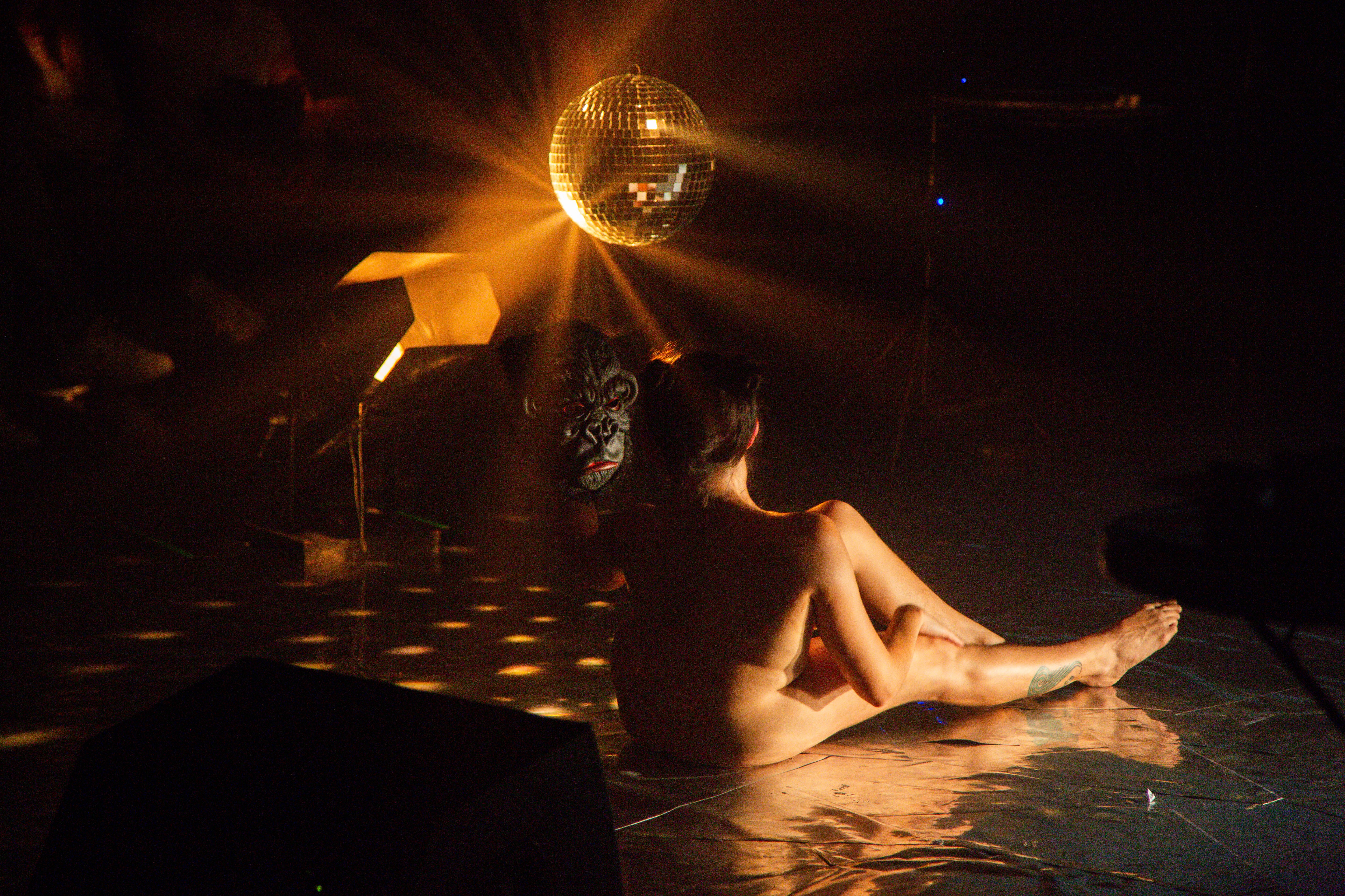 eine Person sitzt nackt mit dem Rücken zur Kamera auf einem silbrigem Boden und hält eine Gorilla Maske. bDas Licht ist warm und eine Diskokugel hängt im Hintergrund.