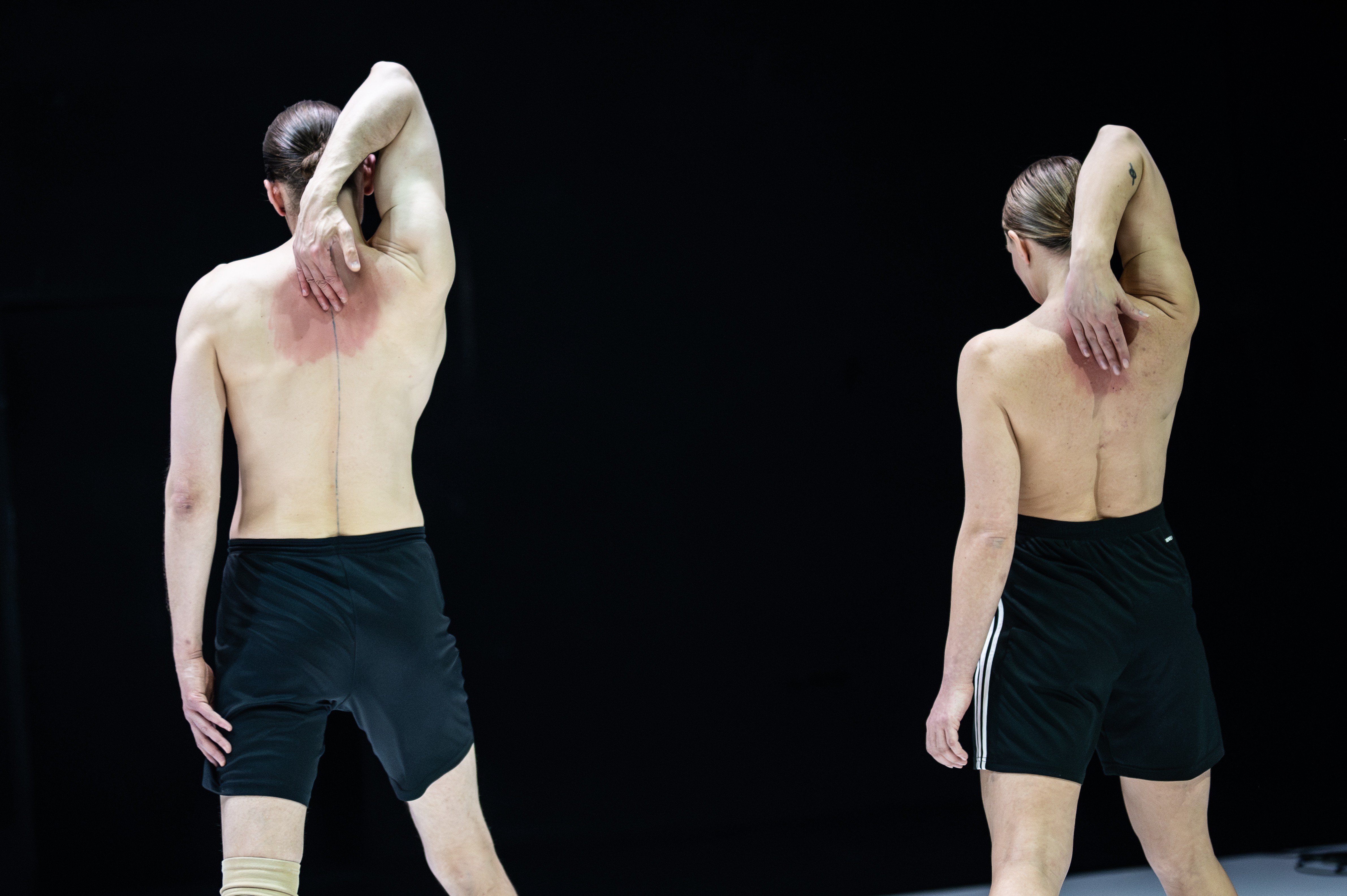 Die beiden Performer*innen stehen mit dem Rücken zum Publikum. Sie sind oberkörperfrei und berühren mit der jeweils rechten Hand ihren Rücken.