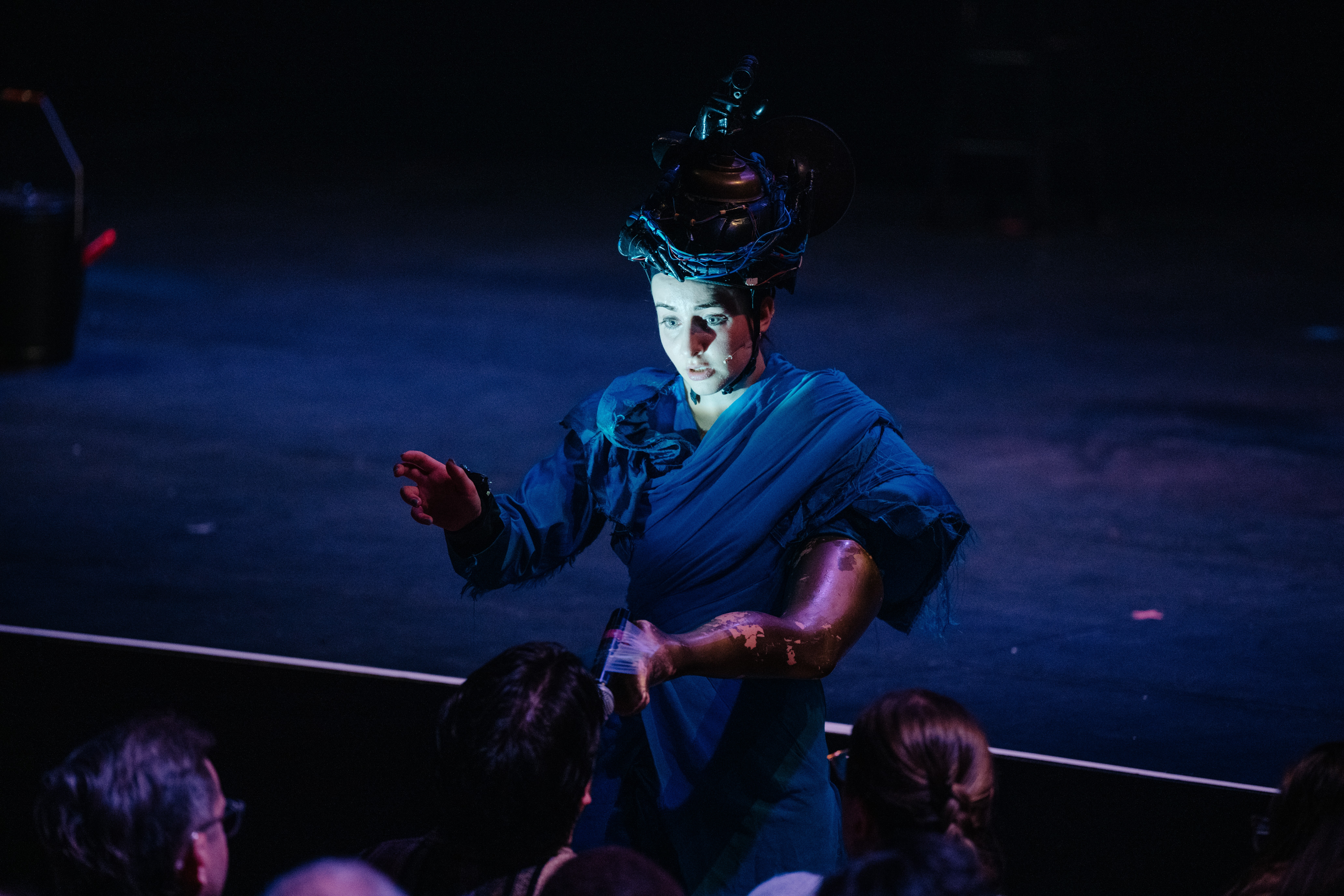 Eine Person spricht zum Publikum. Der Hintergrund ist dunkelblau beleuchtet und ihr Gesicht wir heller beleuchtet. Ein Arm hat sie in einem Puppenbein drin und auf dem Kopf trägt sie einen grossen Apparat.