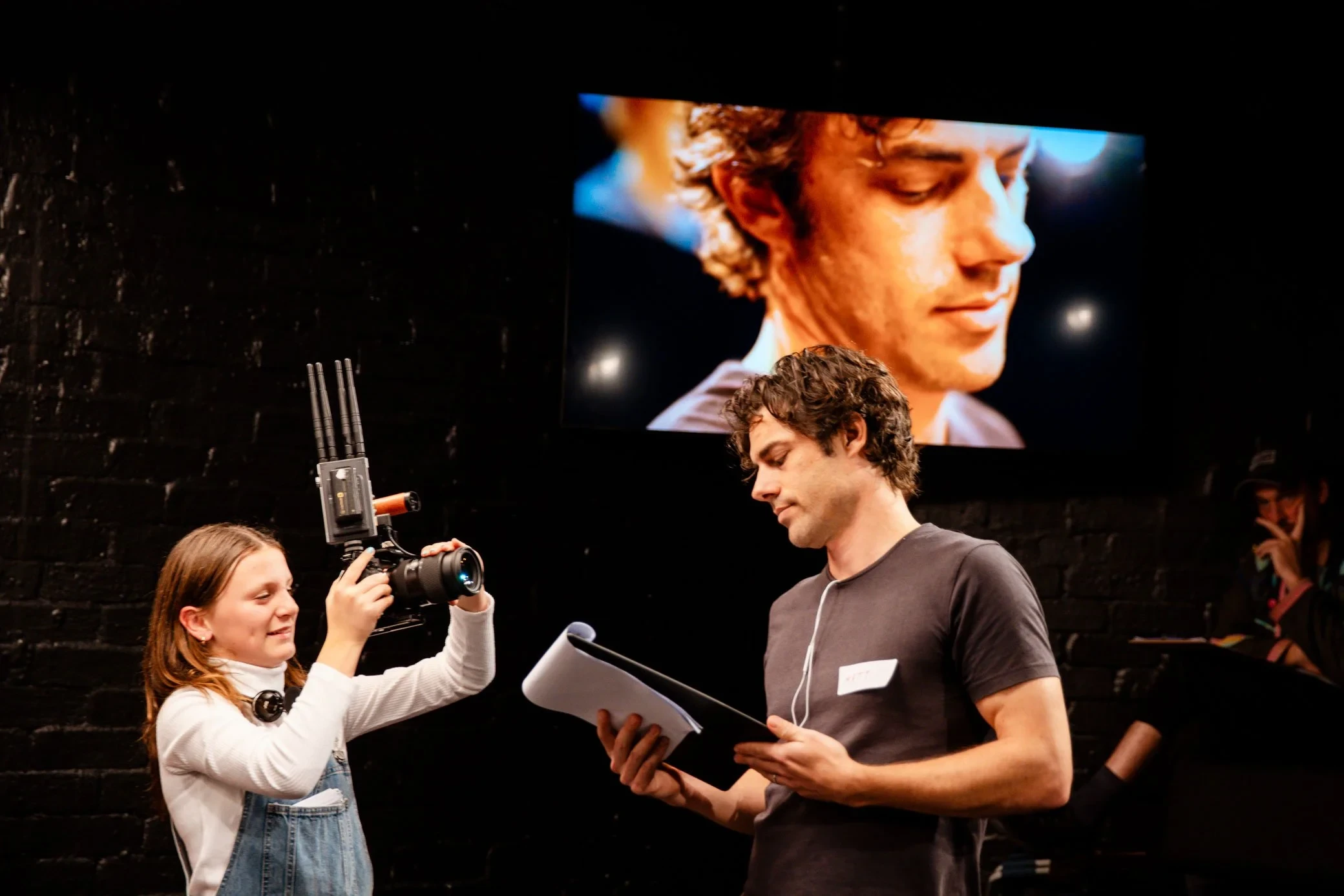 links hält ein Kind eine Kamera und filmt damit eine Person, die konzentriert von einem Skript abliest. Das Gefilmte wird hinter der lesenden Person projiziert.