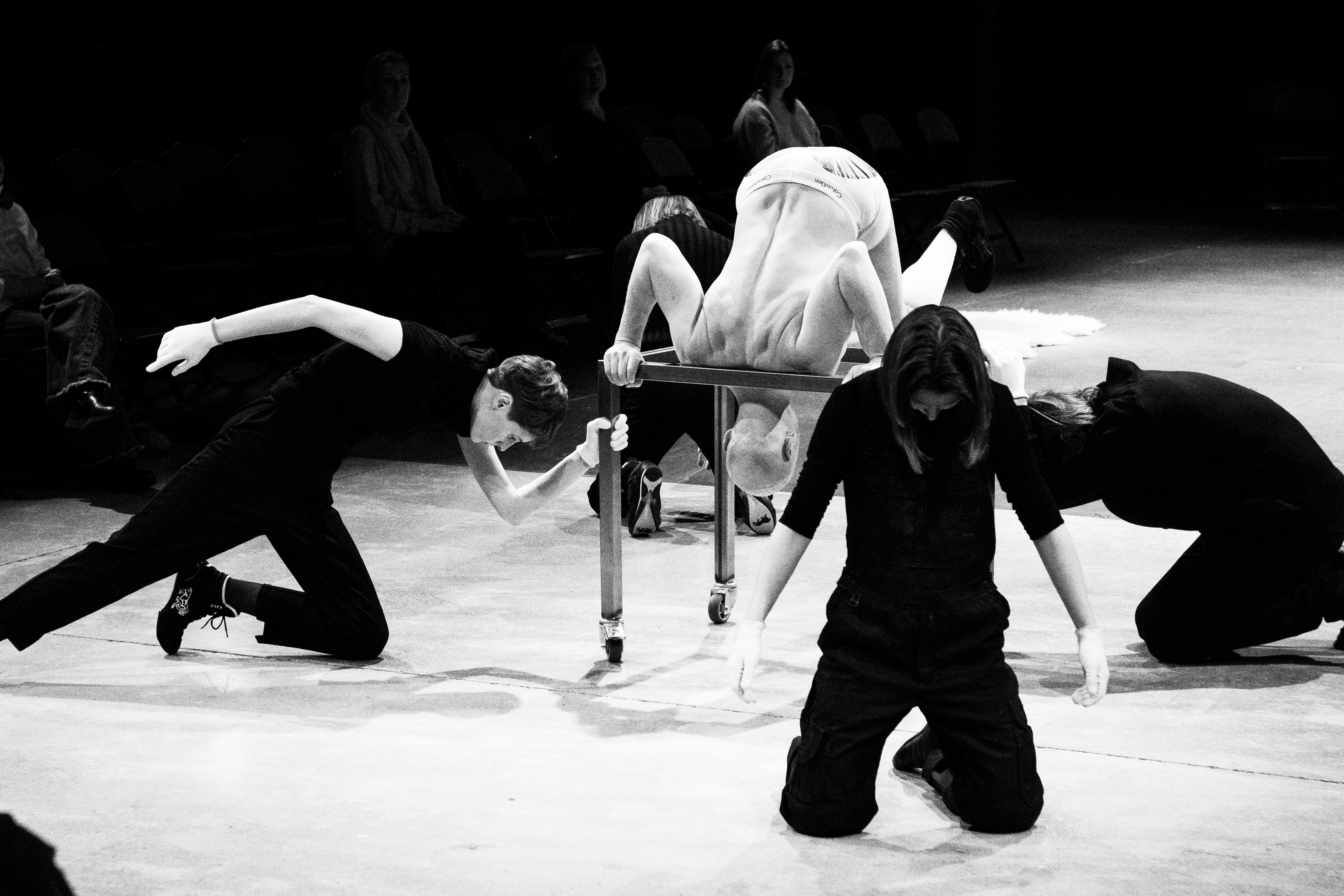 auf einem schwarz-weiss Bild  hocken und bücken sich Personen in verschiedene Richtungen und Positionen gegen Boden. In der Mitte beugt sich eine Person in einen metal Rahmen rein.