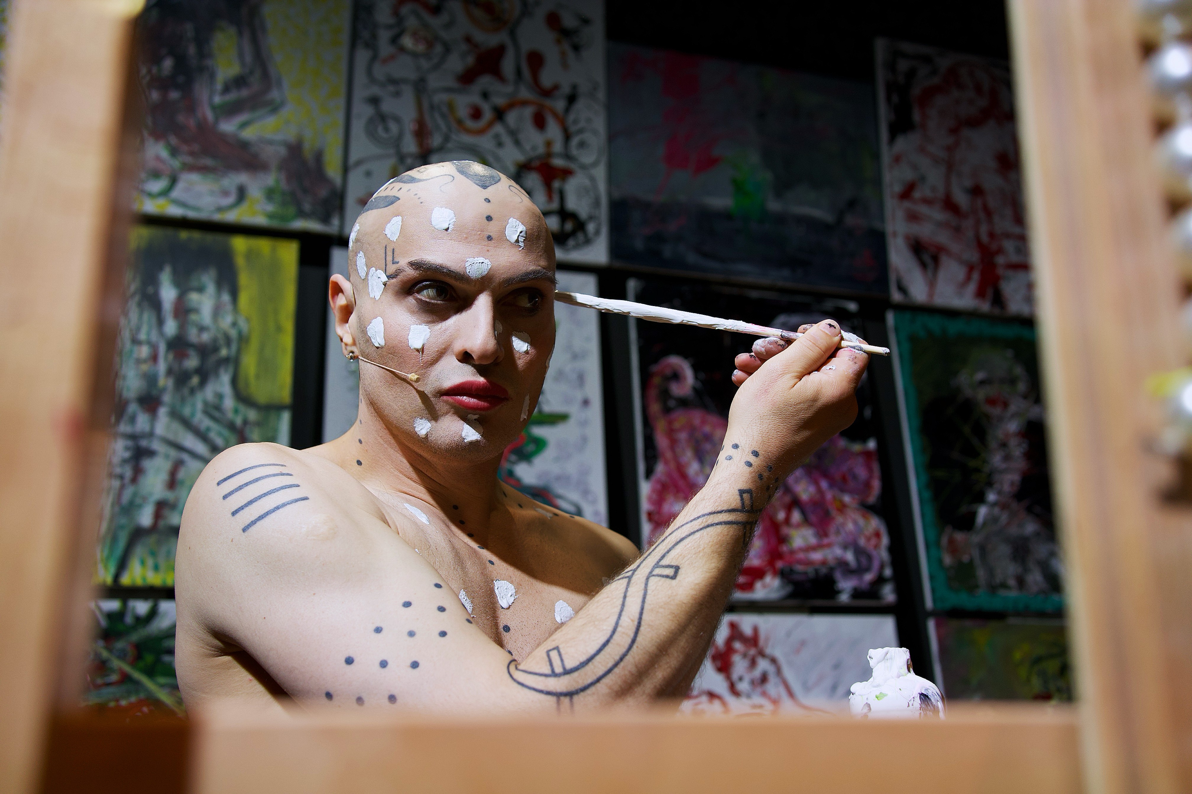 eine Person schminkt sich, die hat am ganzen Körper mit schwarzen Punkten und Linien dekoriert und malt sich gerade weisse grosse Punkte ins Gesicht.