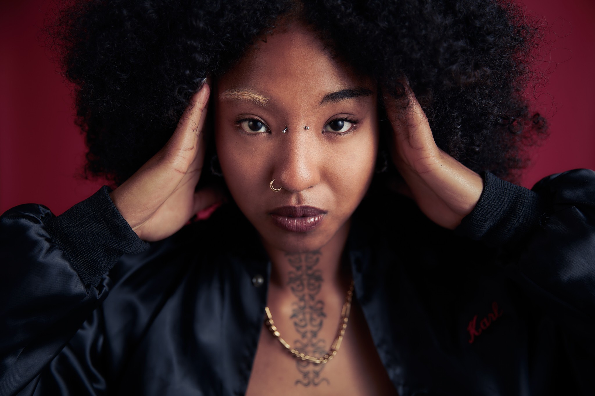 Eine Frau mit Tattoos, Piercings und dunklen Locken, die direkt in die Kamera schaut und dabei ihre Hände an den Kopfseiten abstützt.