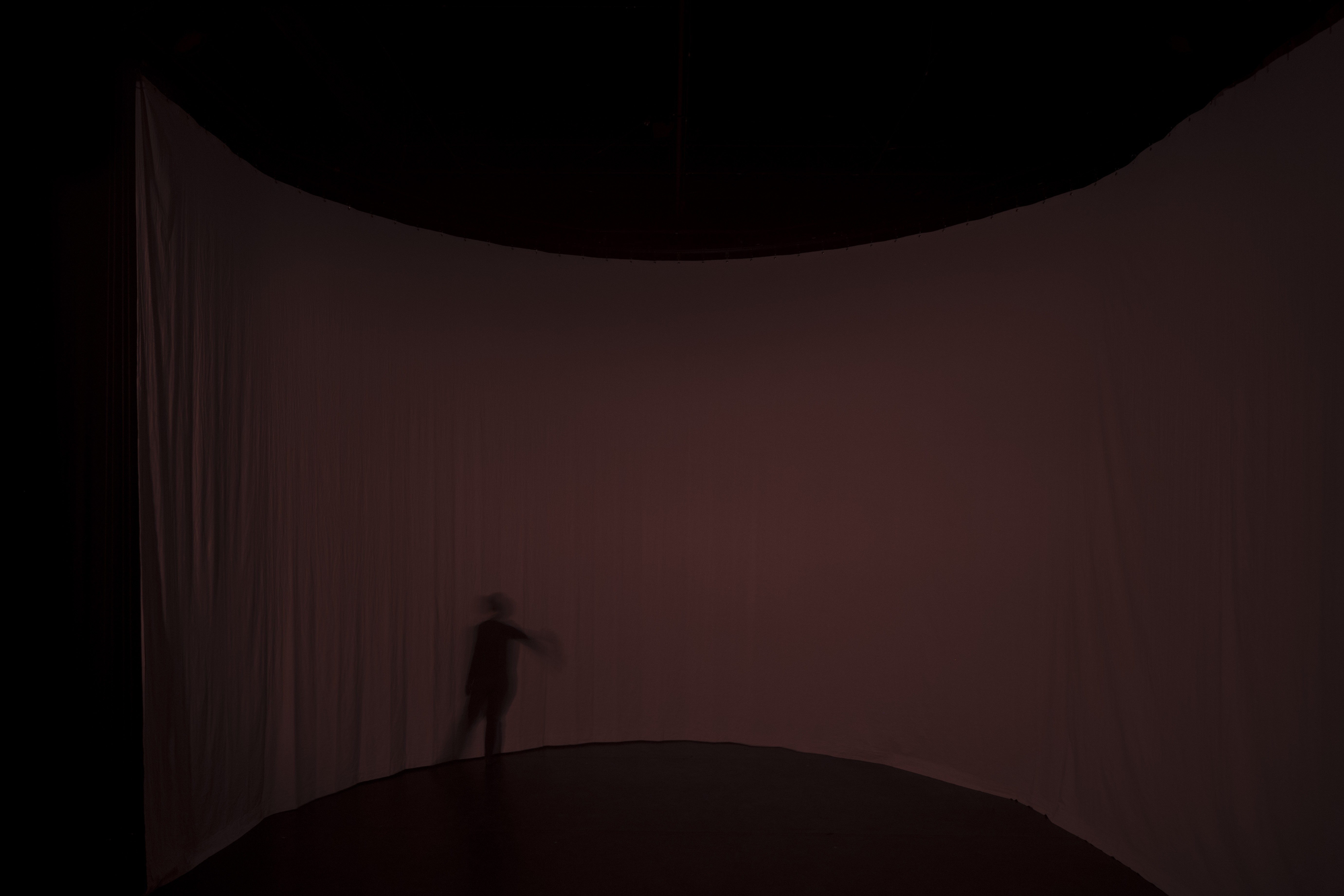 Ein rund gespannter Vorhang in einem roten Dämmerlicht. Ein Schatten einer Person in Bewegung zeichnet sich auf dem Vorhang ab.