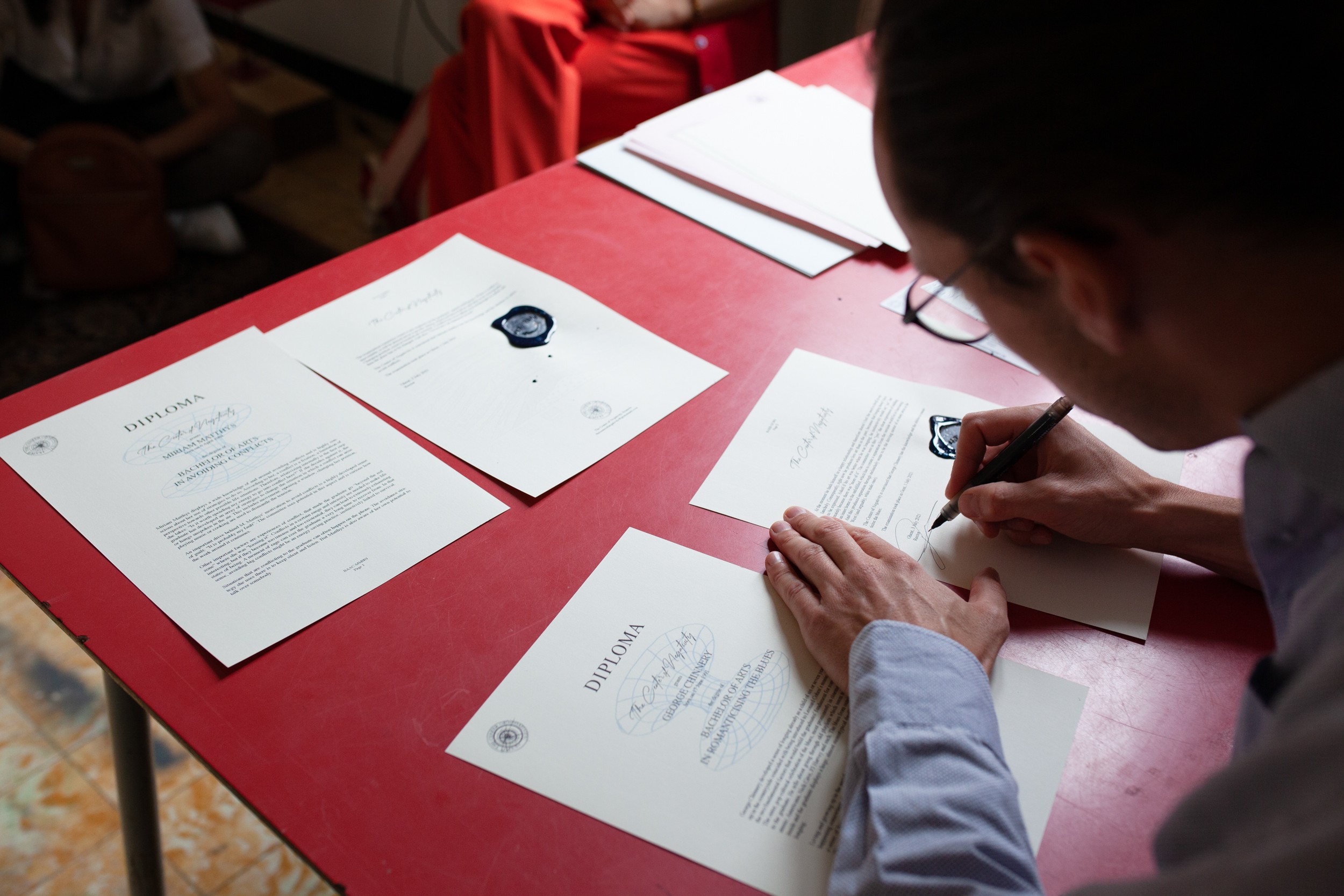 Auf einem roten Tisch liegen verschiedene weisse beschriebene Papiere, einige sind übertitelt mit «Diploma». Rechts im Bild sieht man, wie sich eine Person über den Tisch beugt und eines der Papiere unterschreibt.
