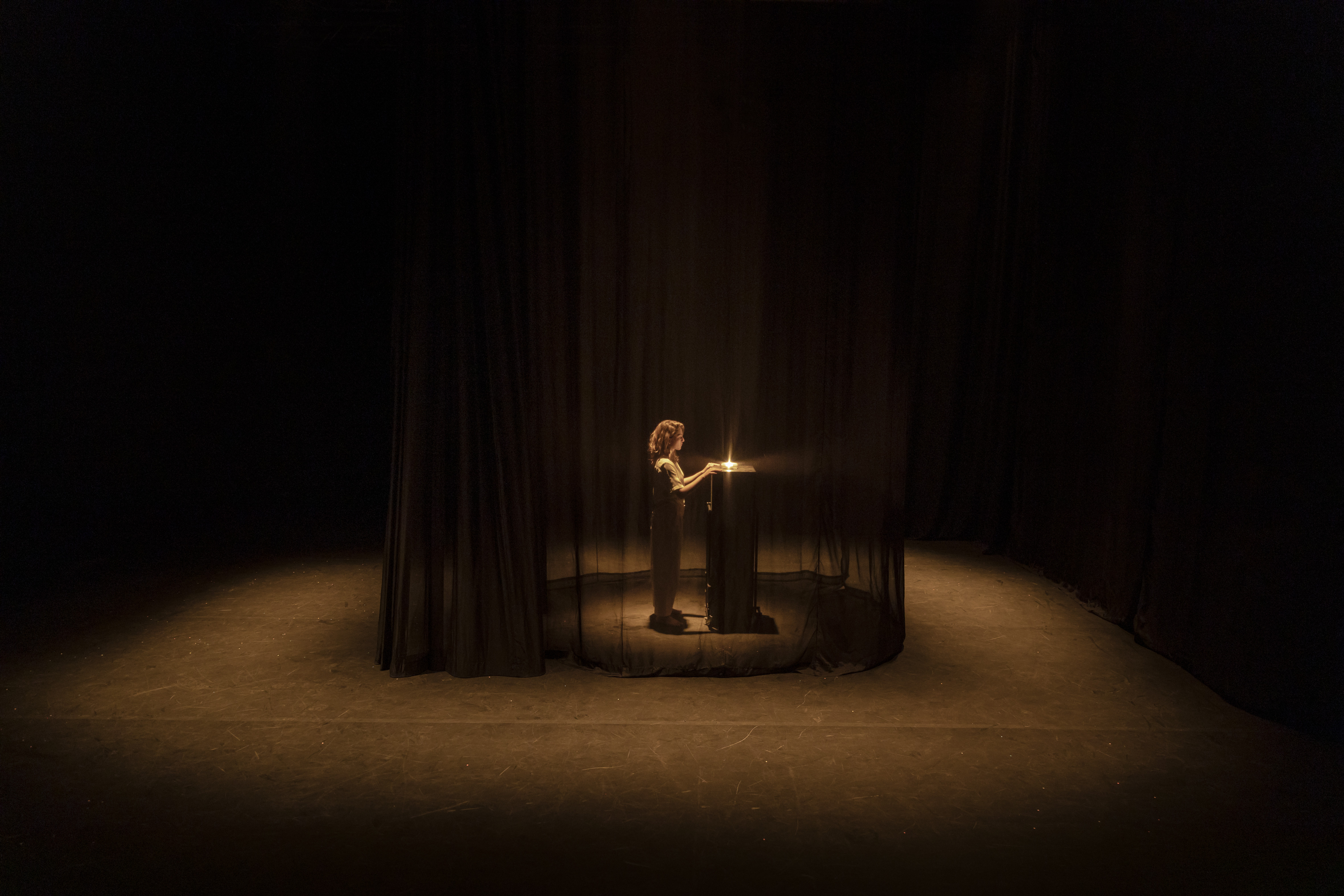 Der abgebildete Raum ist sehr dunkel und nur schwach beleuchtet. In seiner Mitte steht Charlotte De Bruyne vor einem hohen Tisch mit einem kleinen Licht darauf. Charlotte De Bruyne ist umgeben von einem transparenten schwarzen Vorhang.