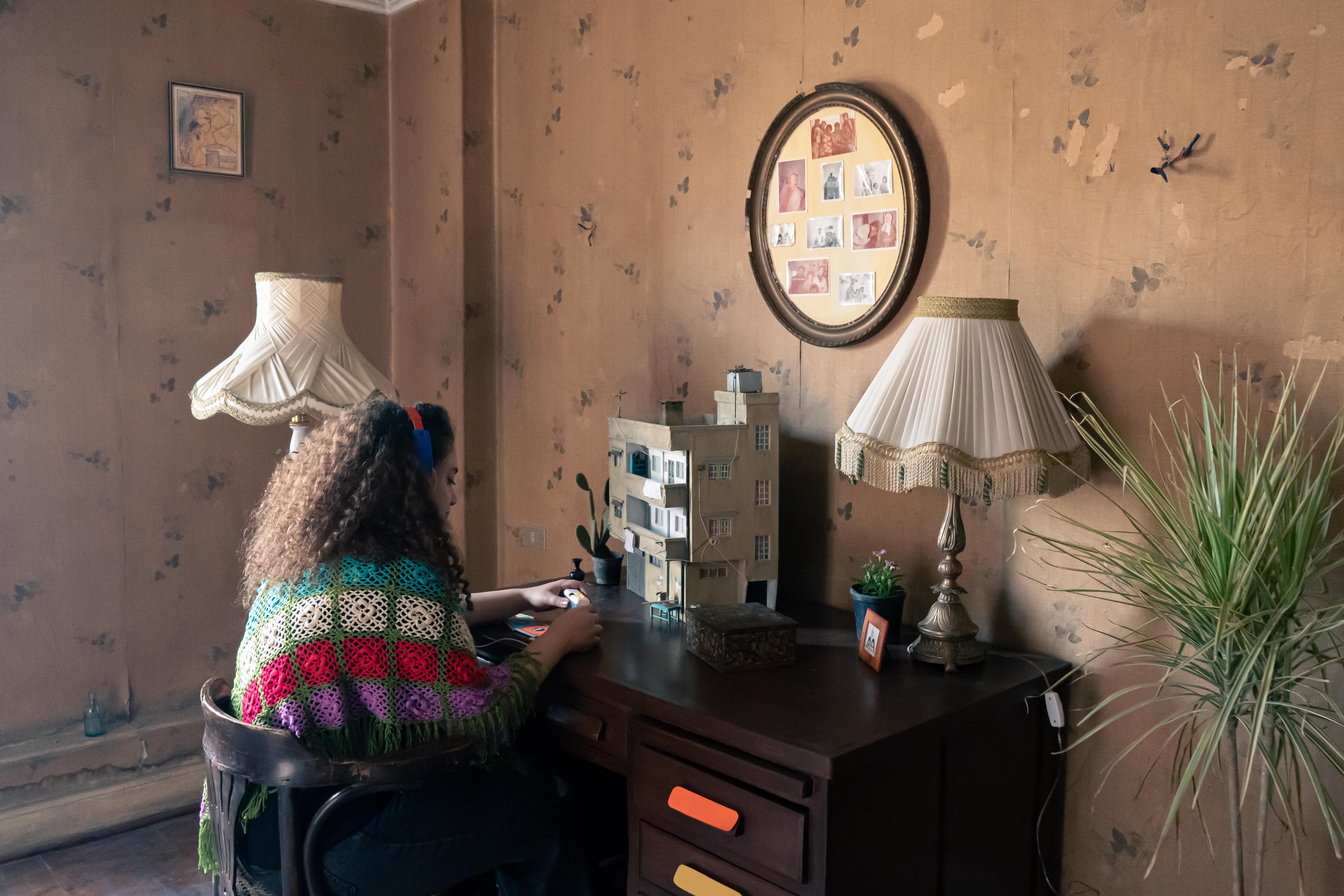 Das Bild zeigt ein gemütliches Zimmer mit brauner Tapete. Eine Person mit Kopfhörern sitzt an einem Schreibtisch. Dort steht das Modell eines Hauses und darüber hängen viele Familienfotos.