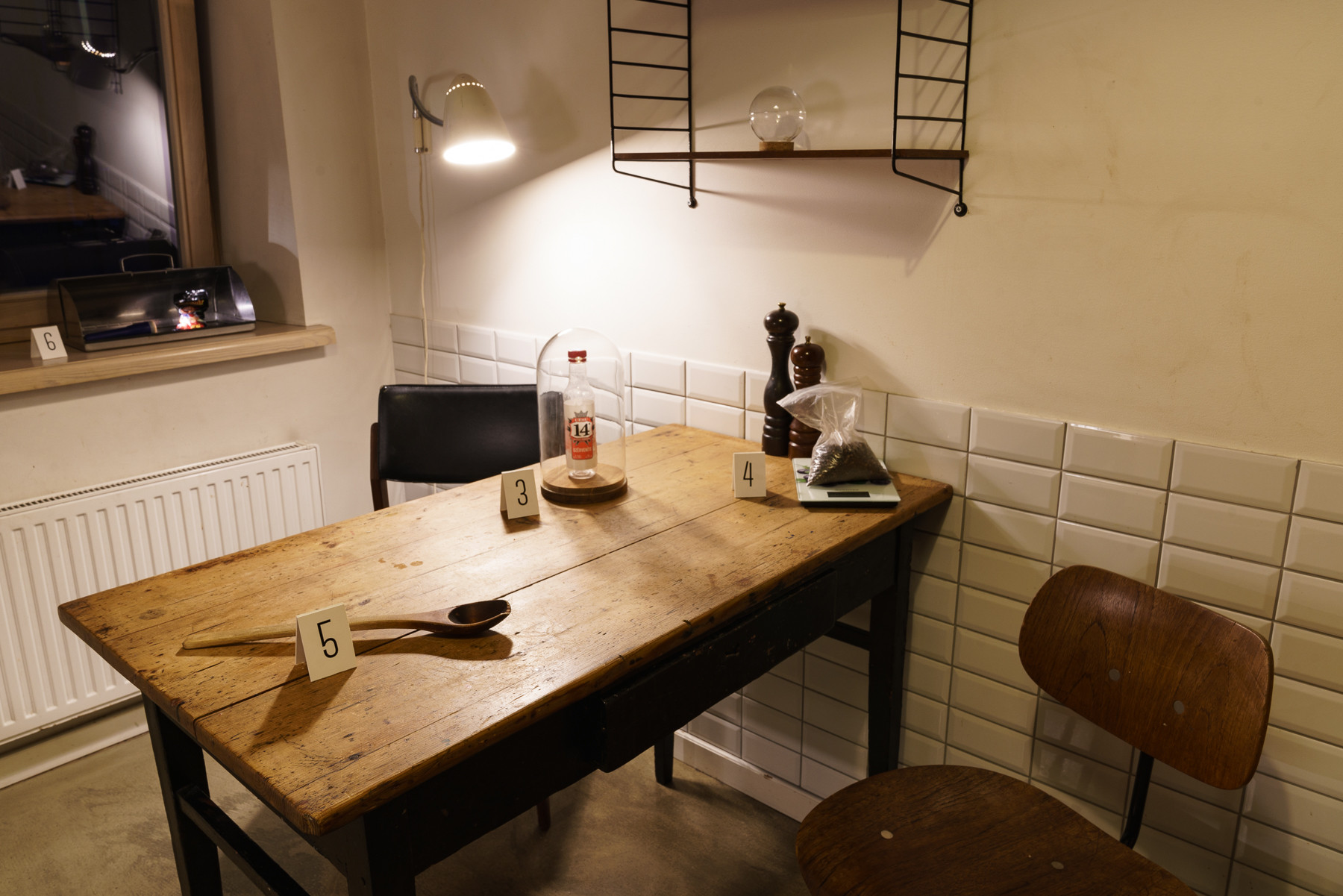 Das Bild zeigt eine Küche mit einem Holztisch und zwei Stühlen. Auf dem Tisch befinden sich ein Kochlöffel und zwei Pfeffermühlen, die je ein Schild mit einer Nummer drauf haben.