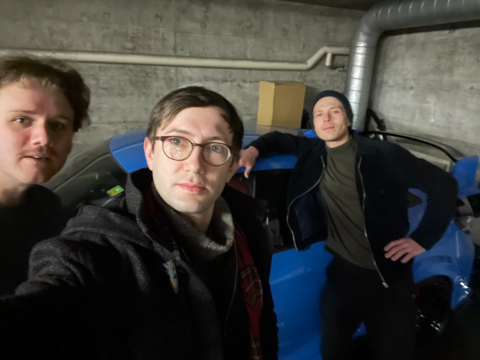 Der Raum ist eine Garage, im Hintergrund ein blaues Auto. Drei Personen schauen in die Kamera.