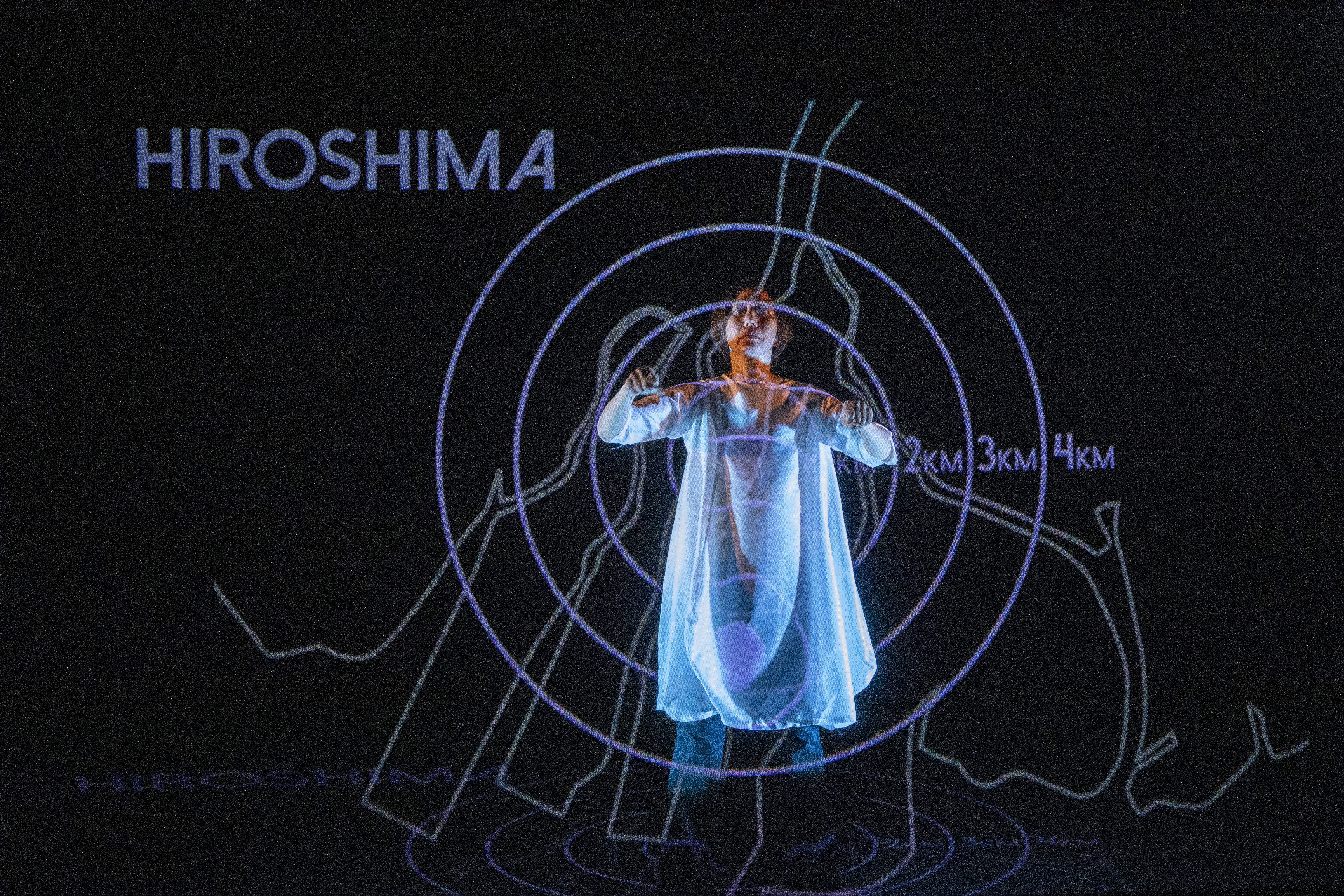 Man sieht eine Person in weissem Kleid, die Armbewegungen macht. Zusätzlich sieht man eine Projektion mit verschiedenen Linien und dem Wort 'Hiroshima'