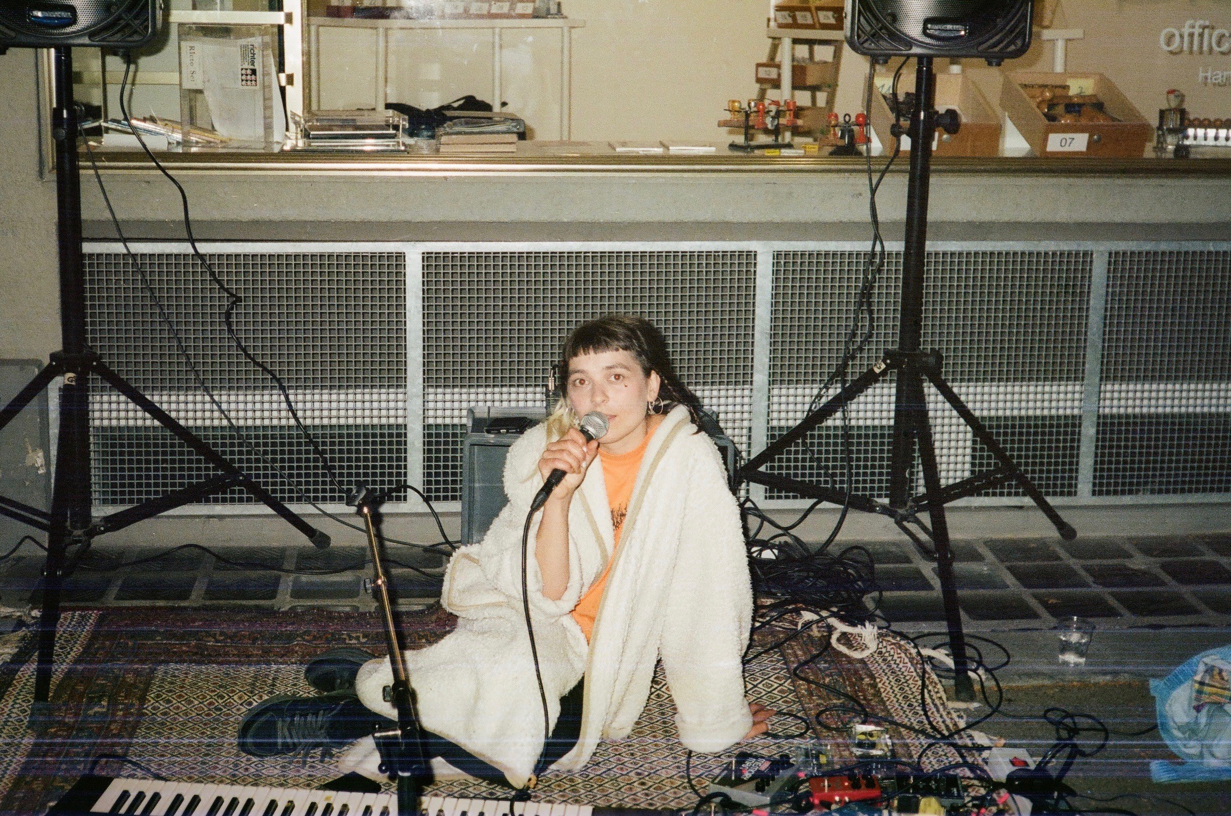 Eine junge Person sitzt in einem weissen Wollmantel am Boden und spricht in ein Mikrofon. Im Raum hat es einen Teppich, Lautsprecher, ein Keyboard und viele Kabel.