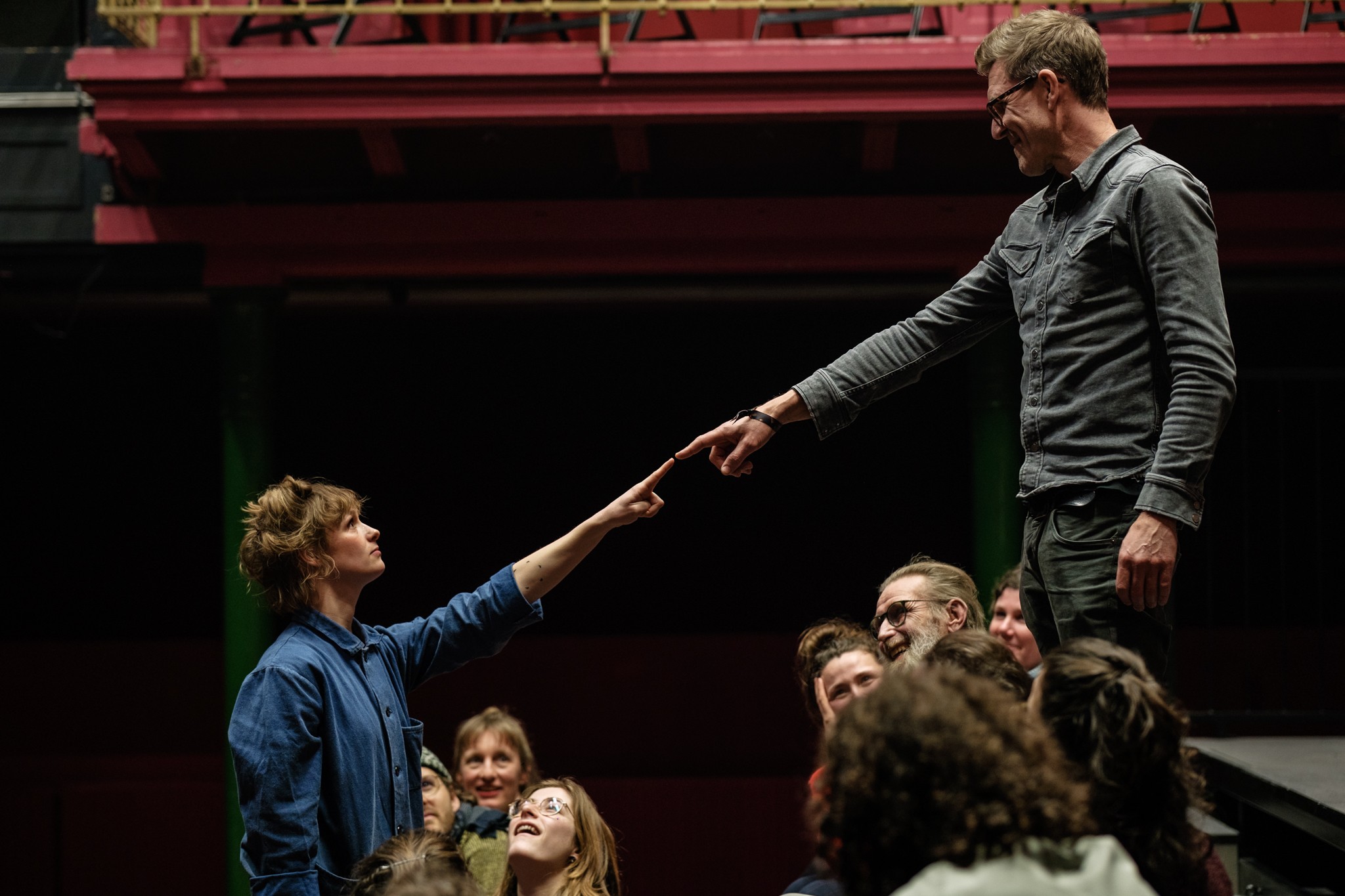 Man sieht auf eine Theatertribüne. Zwei Personen strecken ihren Arm so aus, dass sich ihre Zeigfinger berühren. Die anderen Menschen schauen ihnen dabei zu.