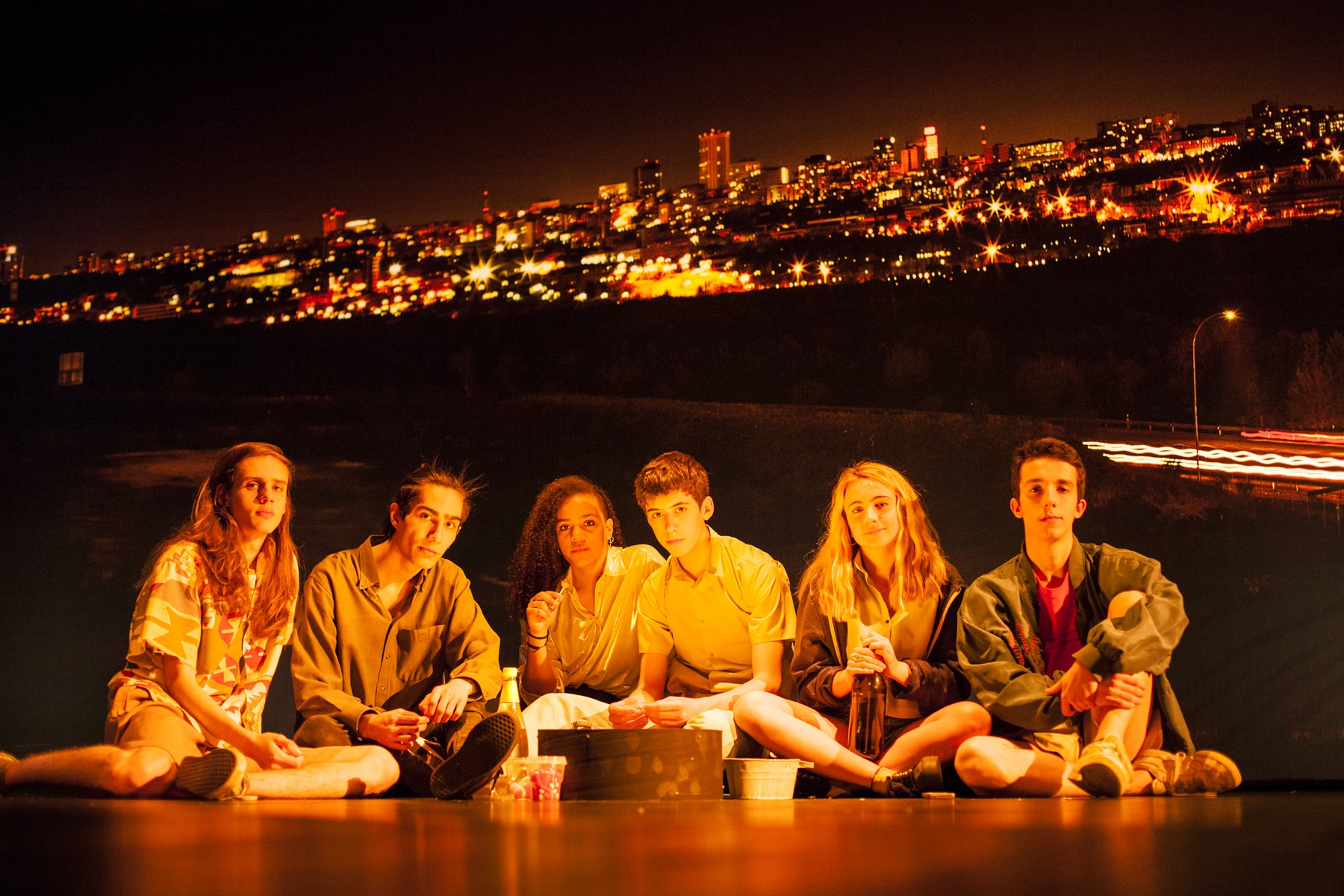 Sechs junge Menschen sitzen am Boden und blicken in die Kamera. Im Hintergrund sieht man eine in der Nacht erleuchtete Stadt.