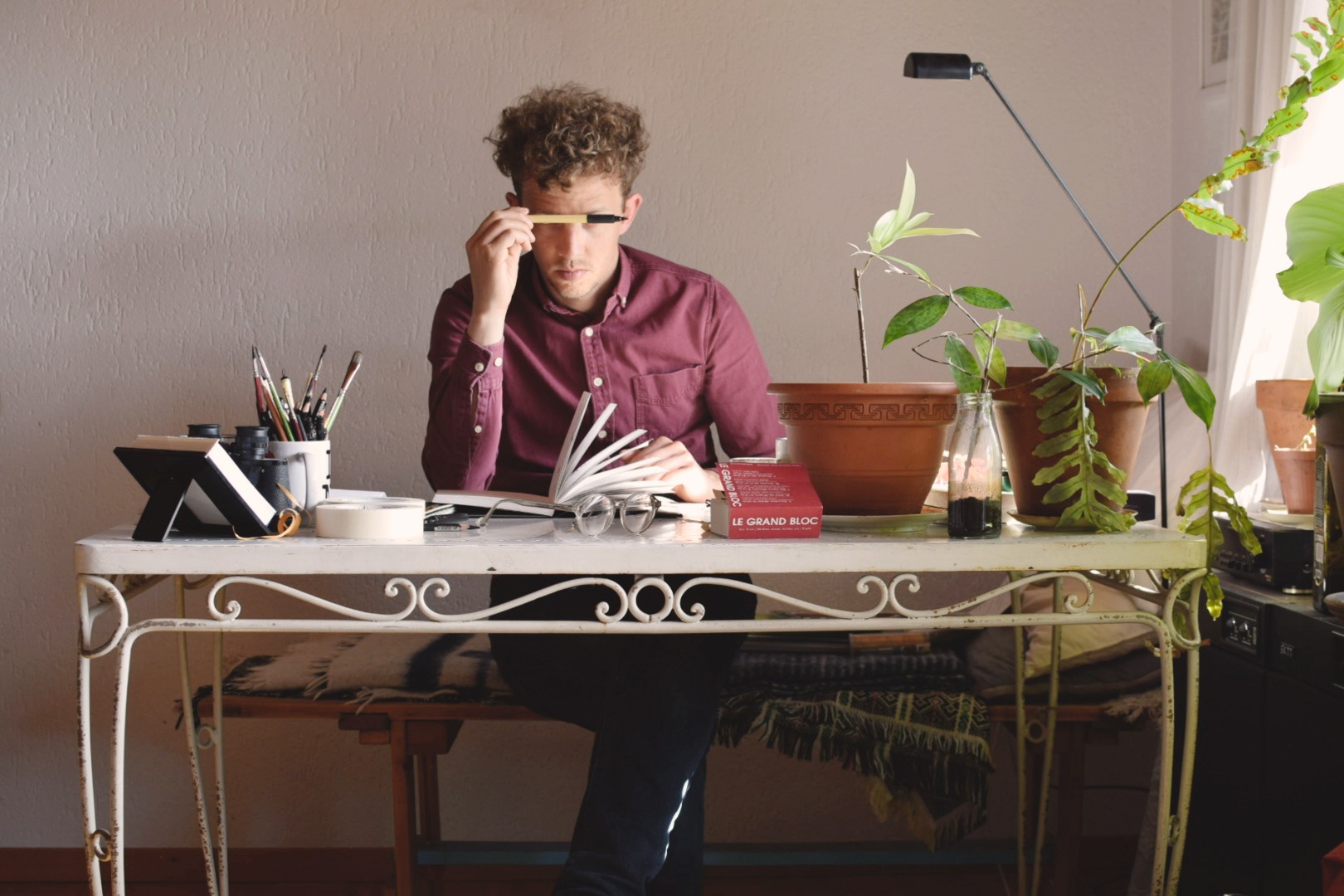 Ein Mann mit Hemd und krausem Haar sitzt an einem Arbeitstisch und hält sich einen Stift vor das Gesicht. Auf dem Tisch liegen Stifte und ein aufgeschlagenes Buch. Daneben stehen Pflanzen.