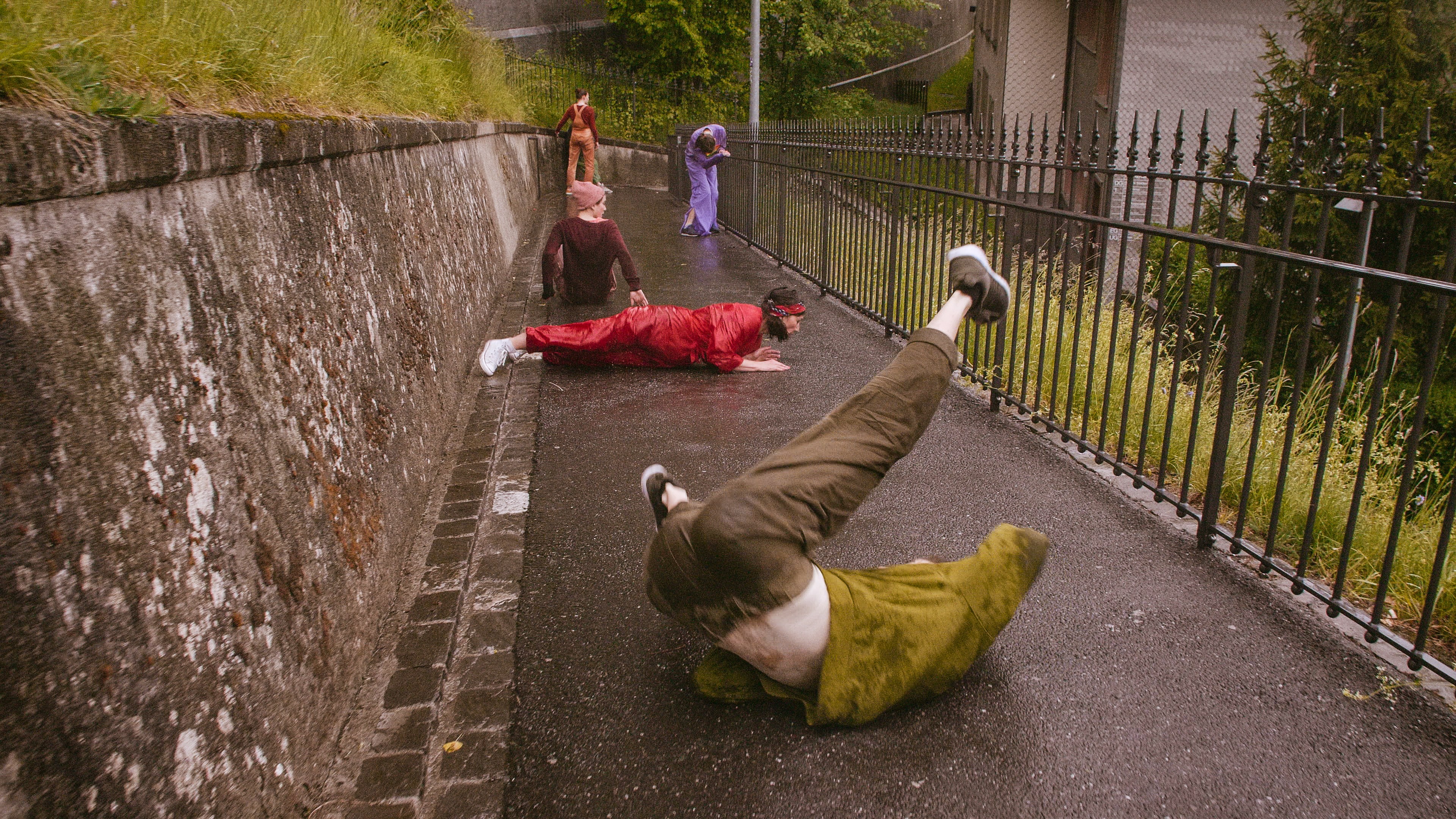 Fünf Menschen in bunten Kostümen bewegen sich tanzend durch eine schmale Strasse. Links von der Strasse eine Mauer und Gras, rechts ein Zaun und Gras.