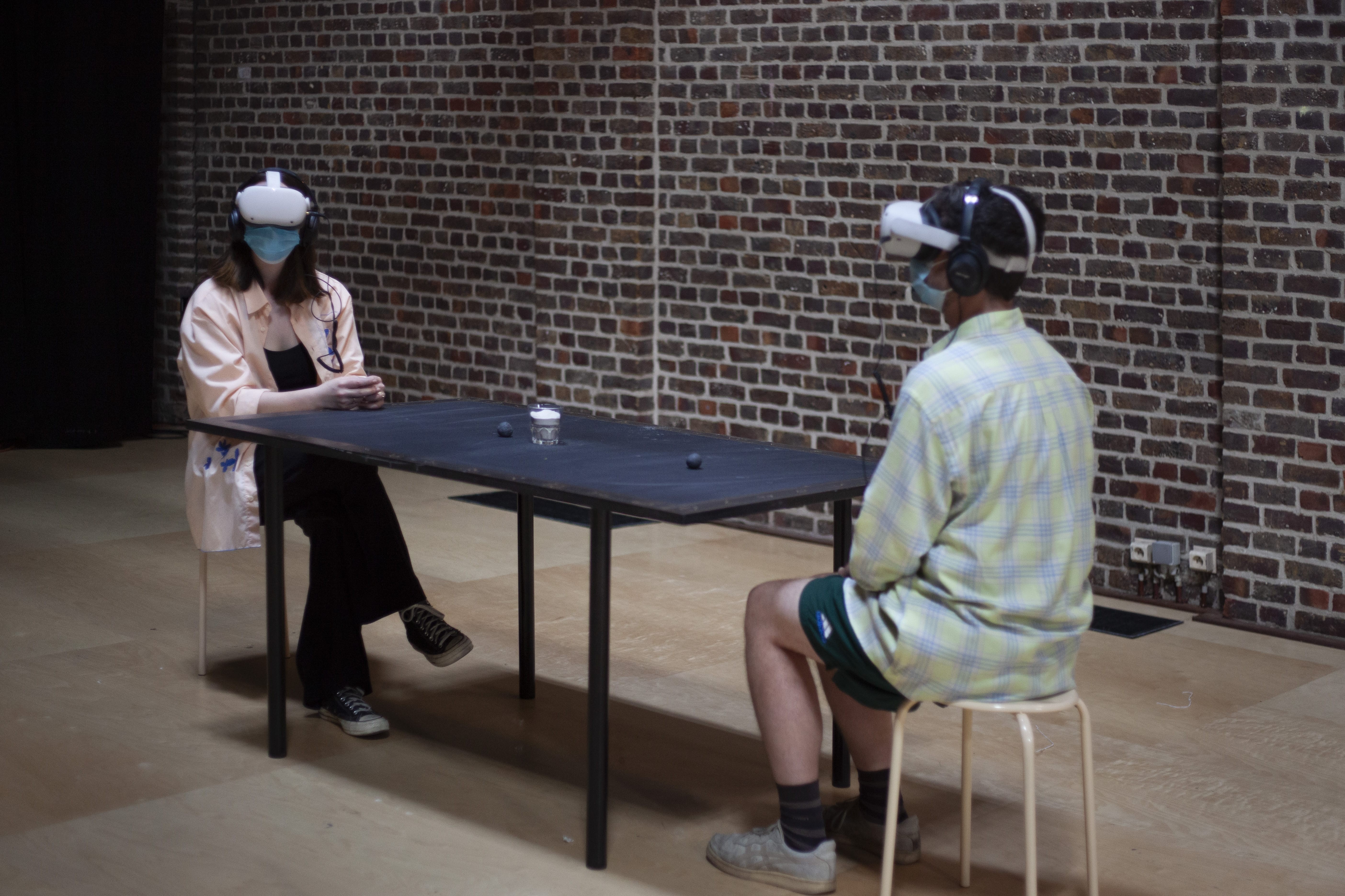 Im Hintergrund eine Backsteinwand. Im Vordergrund sitzen sich zwei Personen an einem schwarzen Tisch gegenüber. Sie haben beide eine VR-Brille auf.